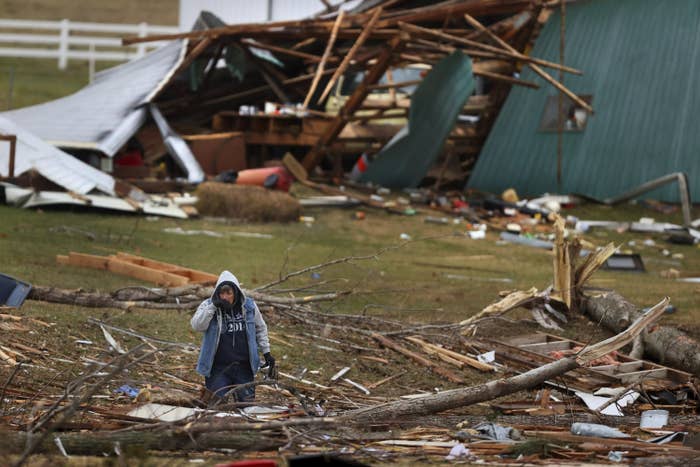 A woman wearing a hooded jacket stands amid debris and fallen tree branches