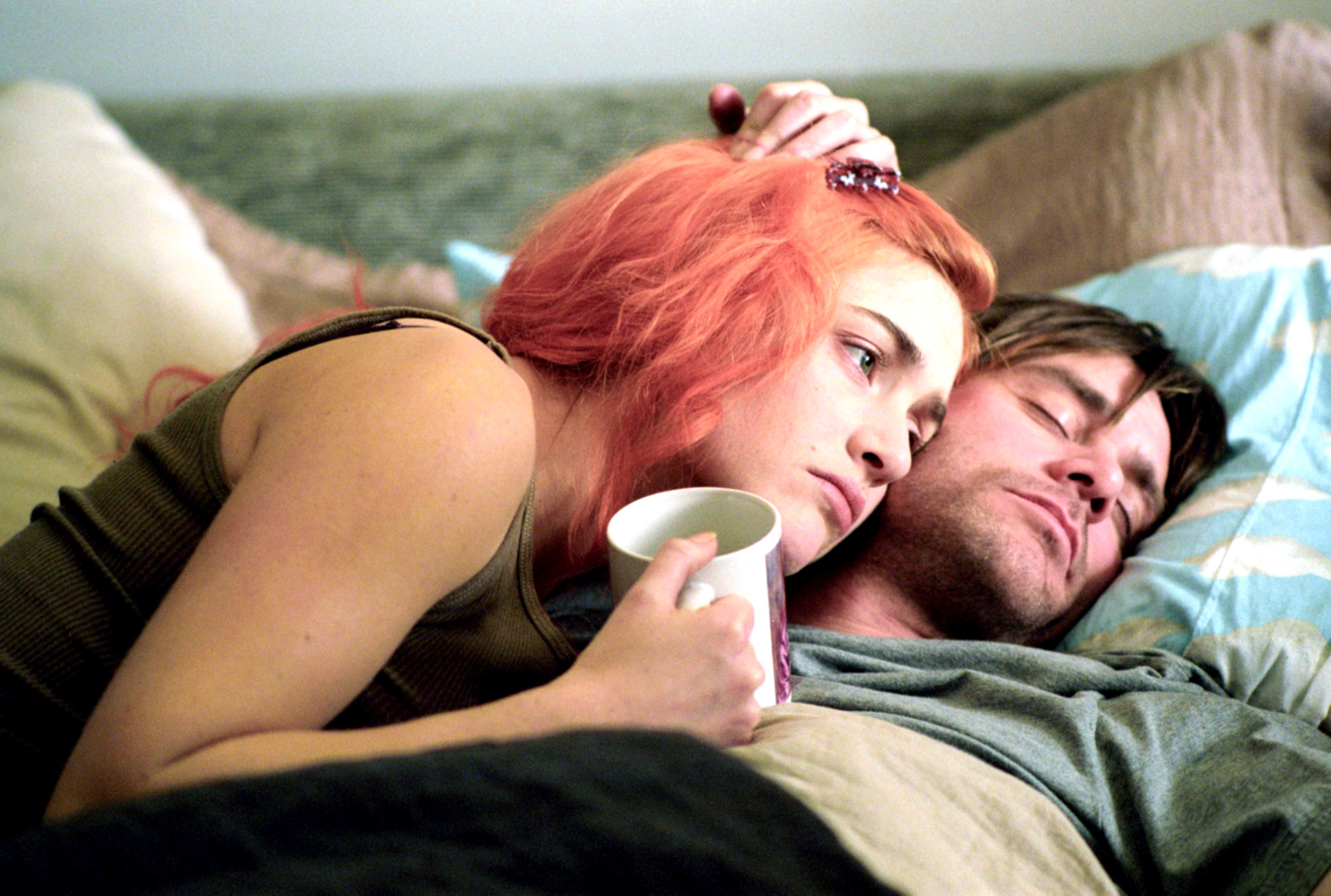 Kate Winslet with dyed peach colored hair, holding a mug, lying on Jim Carrey&#x27;s chest, as he sleeps in bed