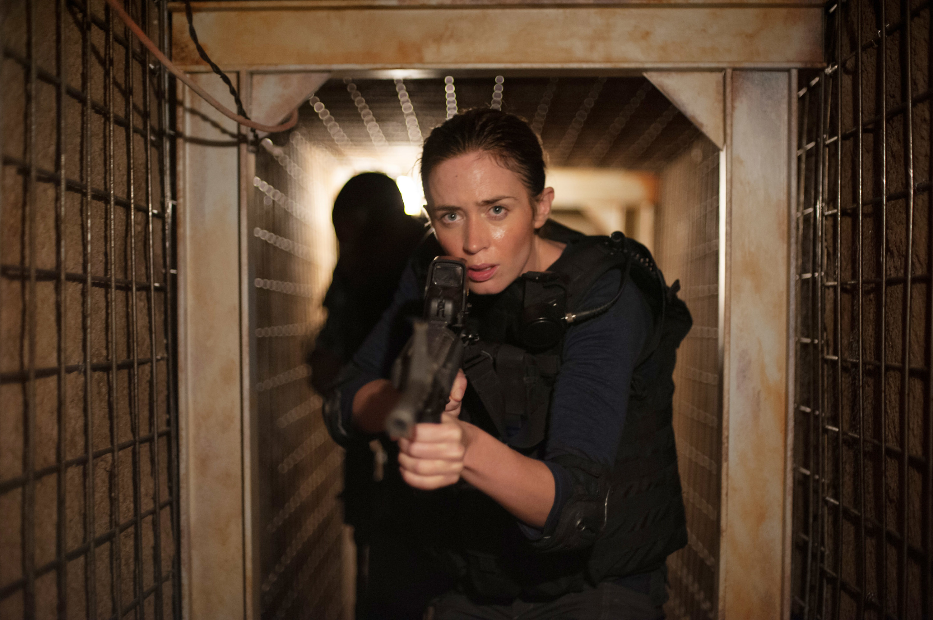 Emily Blunt as an FBI agent, holding a machine gun, wearing a bullet proof vest, walking down a hidden tunnel