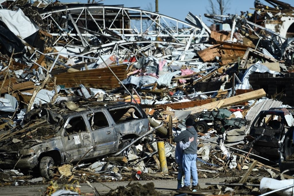 A car with its windows blown out amongst the wreckage as two people look on