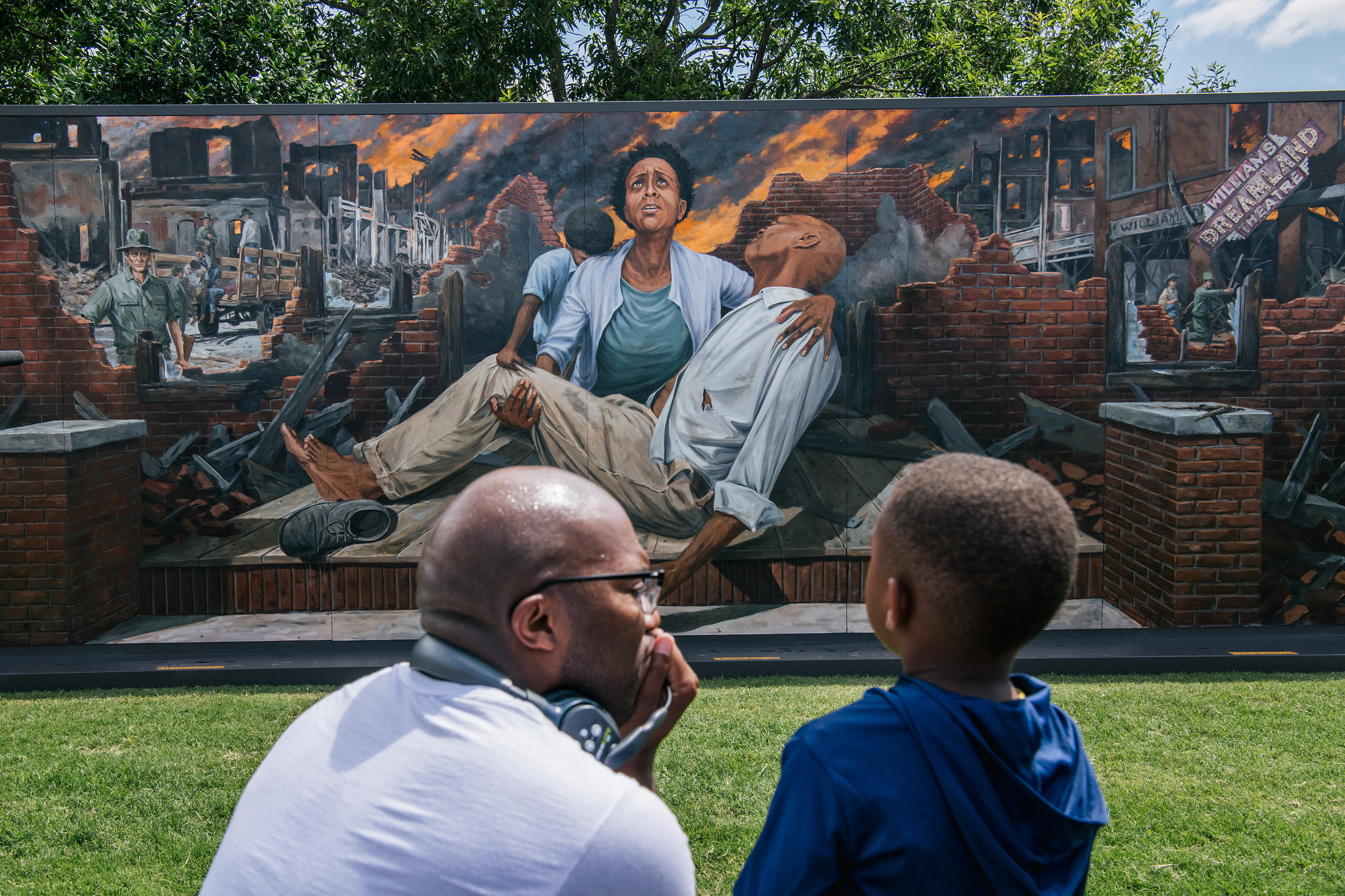 A man in glasses squats on the ground next to a young boy in front of a mural of a woman crying over the body of a man with burning buildings behind him