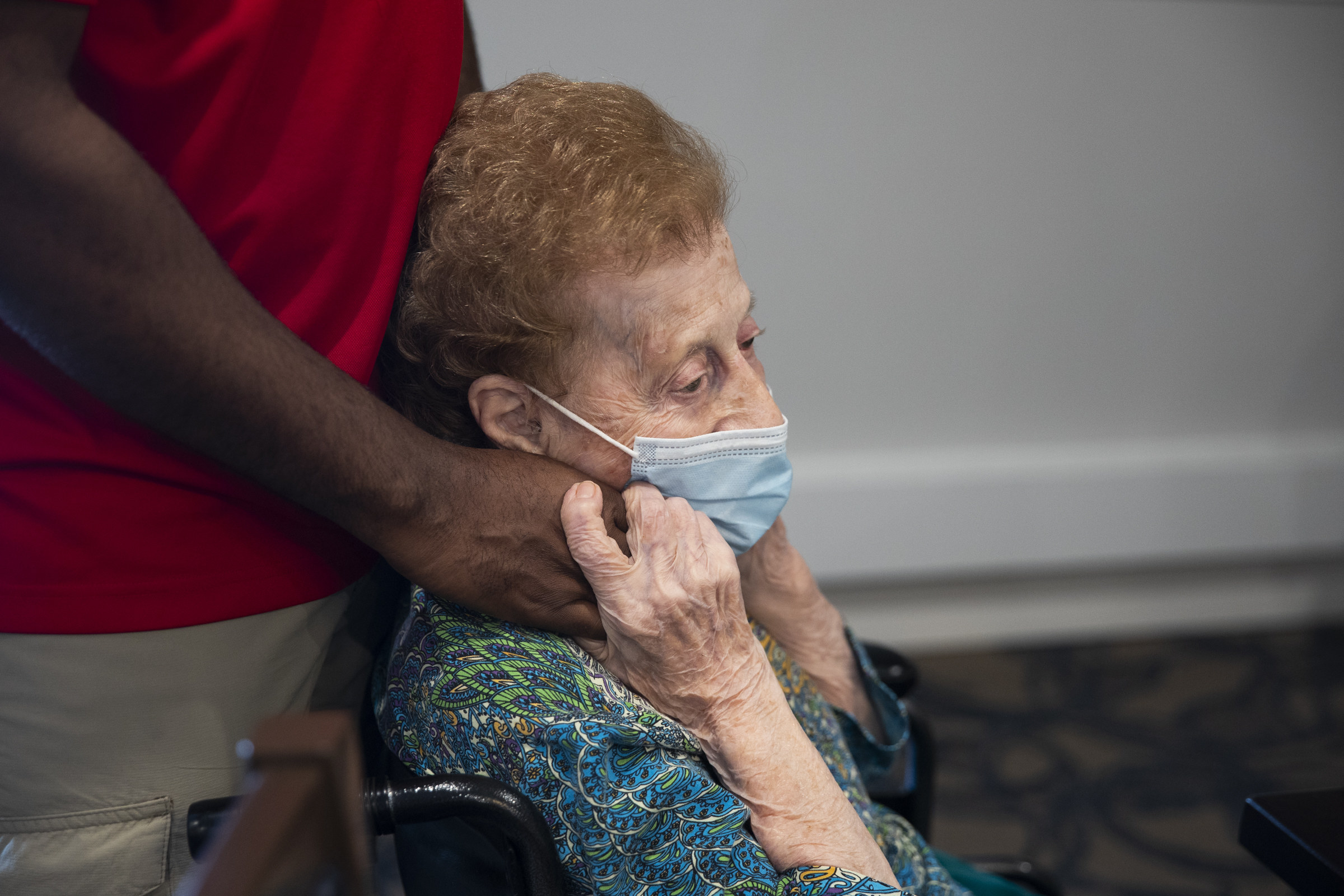 An older woman in a wheelchair grips the hands of a man standing behind her, she is wearing a mask