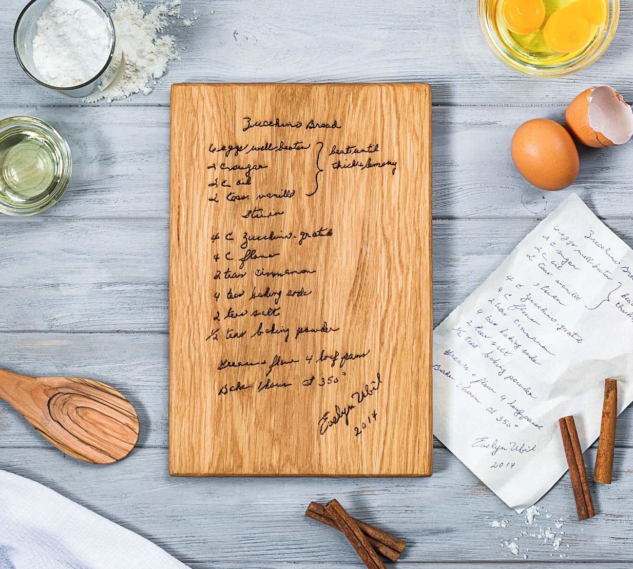 The wooden cutting board engraved with a recipe, next to a piece of paper with the handwritten recipe and surrounded by baking accessories and ingredients
