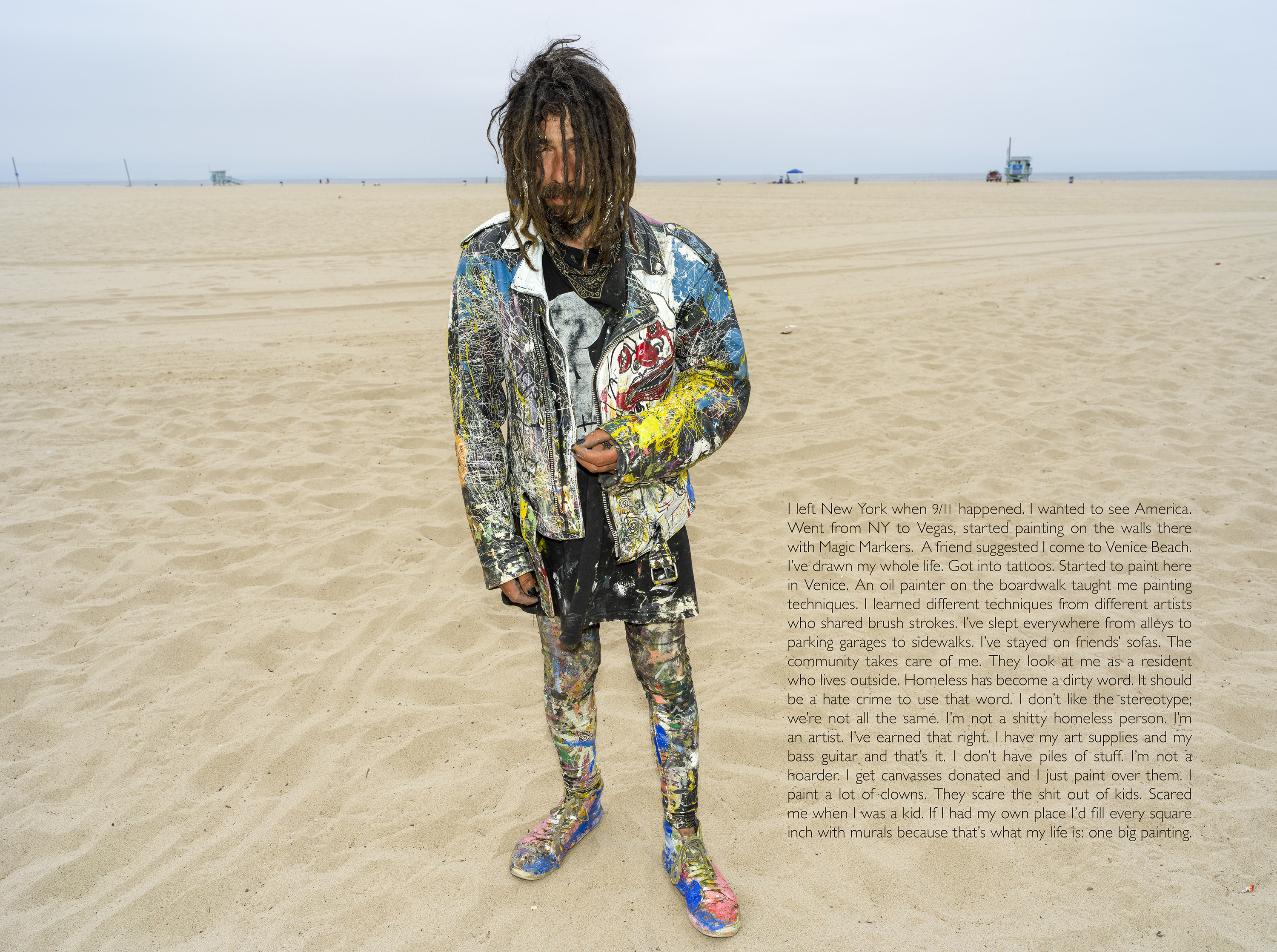 A man wearing paint-splattered clothing stands on a beach