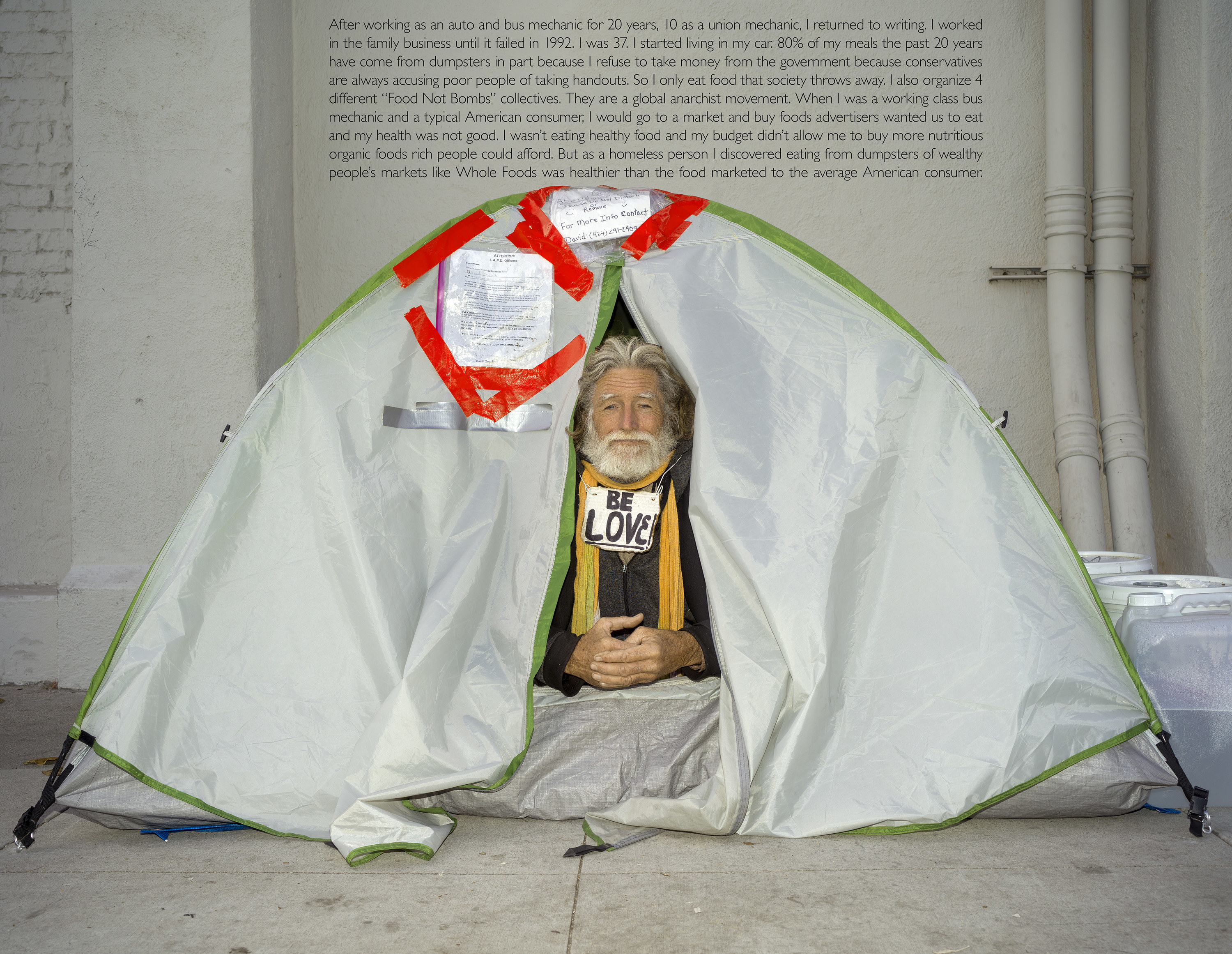 A man sitting in a tent wears a sign that reads &quot;be love&quot;