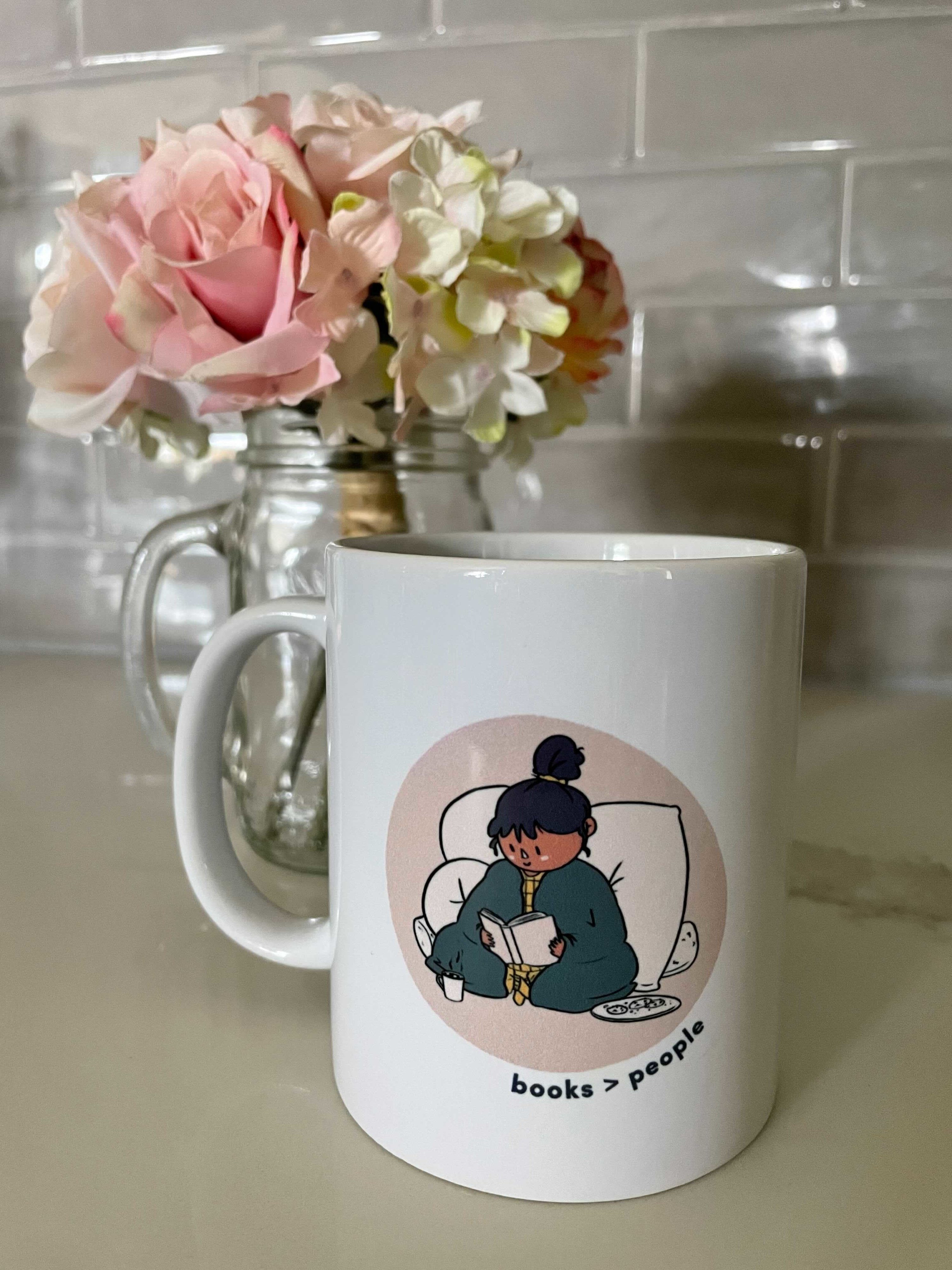 white mug with a person with a messy ponytail leaning against a fluffy pillow with a blanket draped around them reading a book with a mug of cocoa and plate of cookies beside them, text underneath reads books &amp;gt; people