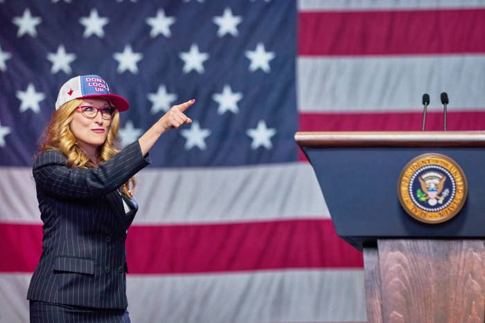 Streep points off-camera while wearing a blazer and baseball hat in front of a podium and American flag