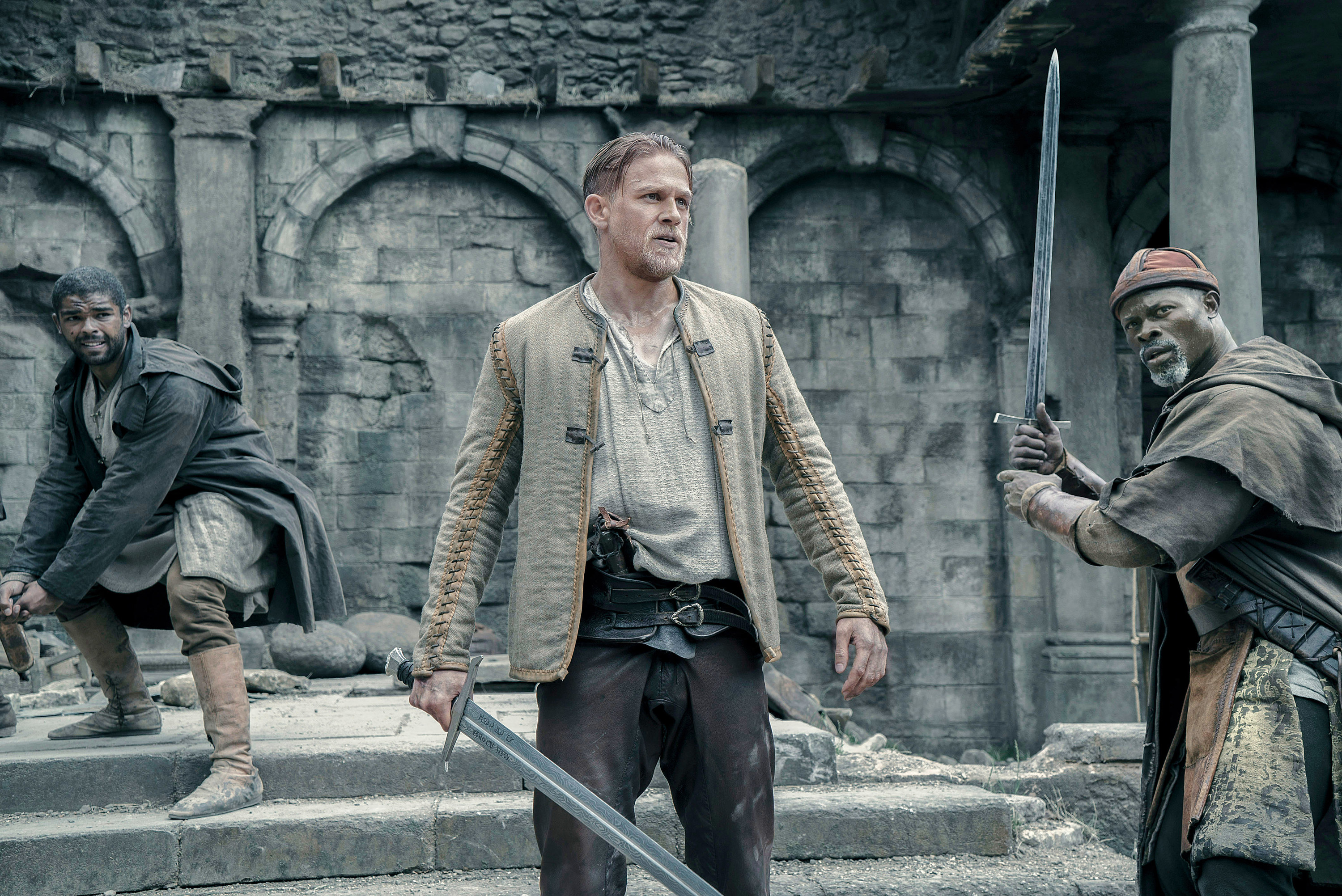 Kingsley Ben-Adir, Charlie Hunnam, and Djimon Hounsou hold swords, like they&#x27;re about to fight, during a scene in the movie