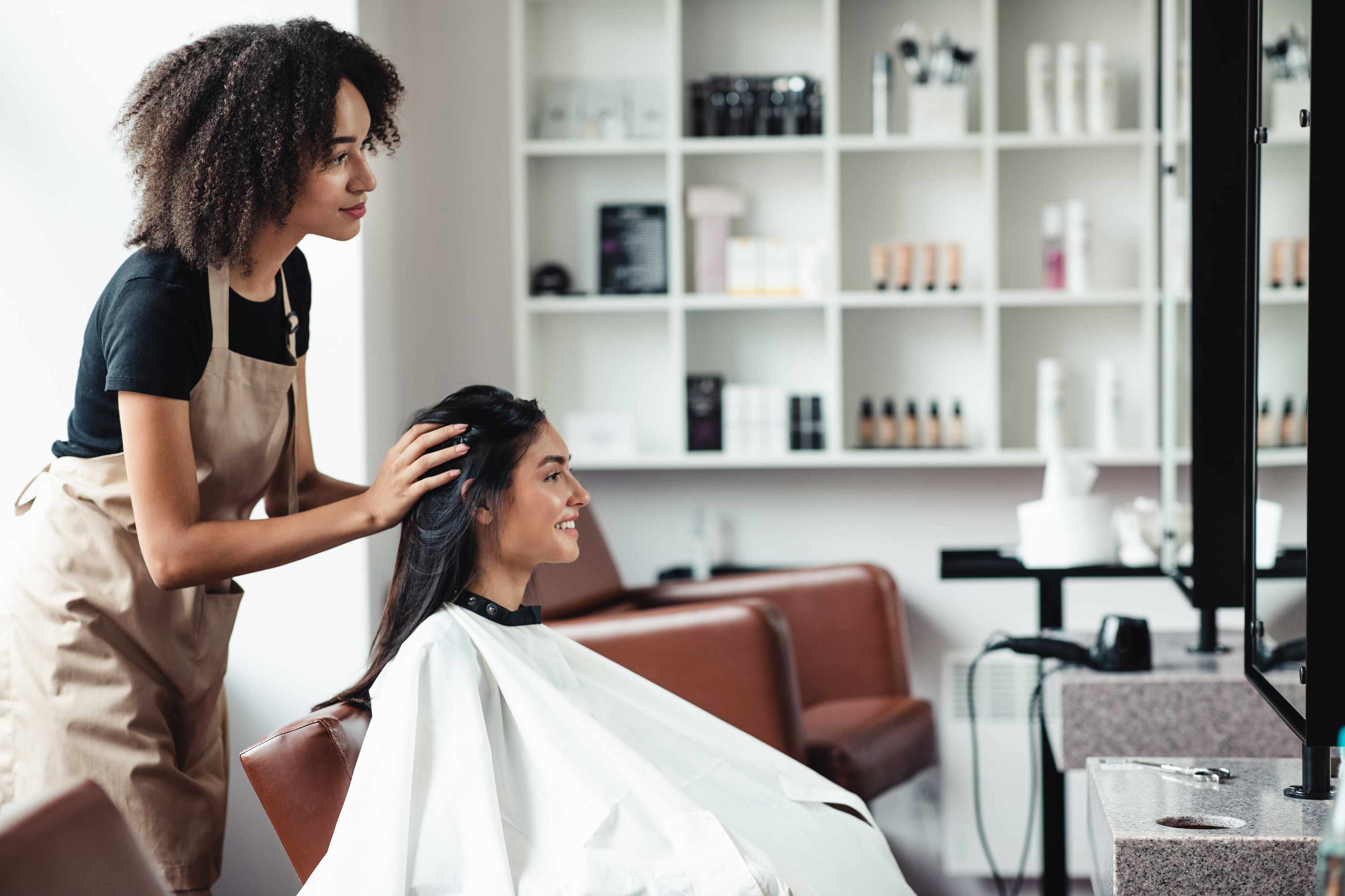 A woman sitting in a hair salon&#x27;s chair with her hairstylist touching her hair