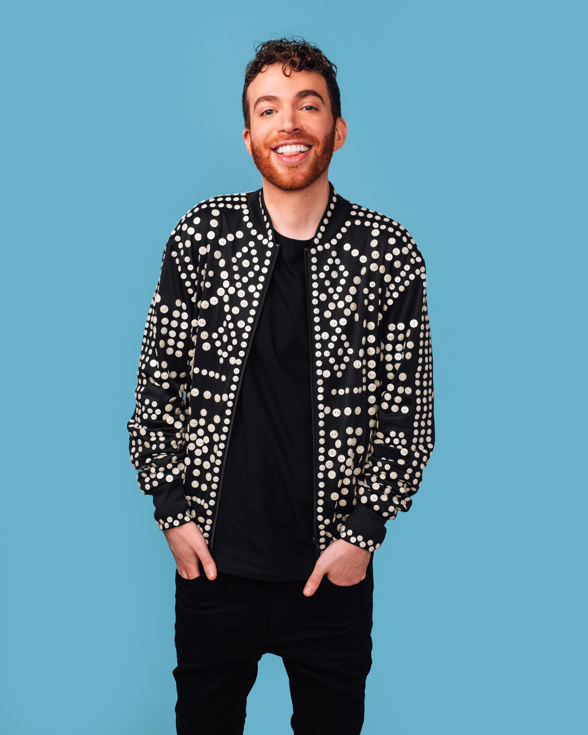 Josh smiles widely as they poses with both hands their pocket and rocks a polka dot jacket