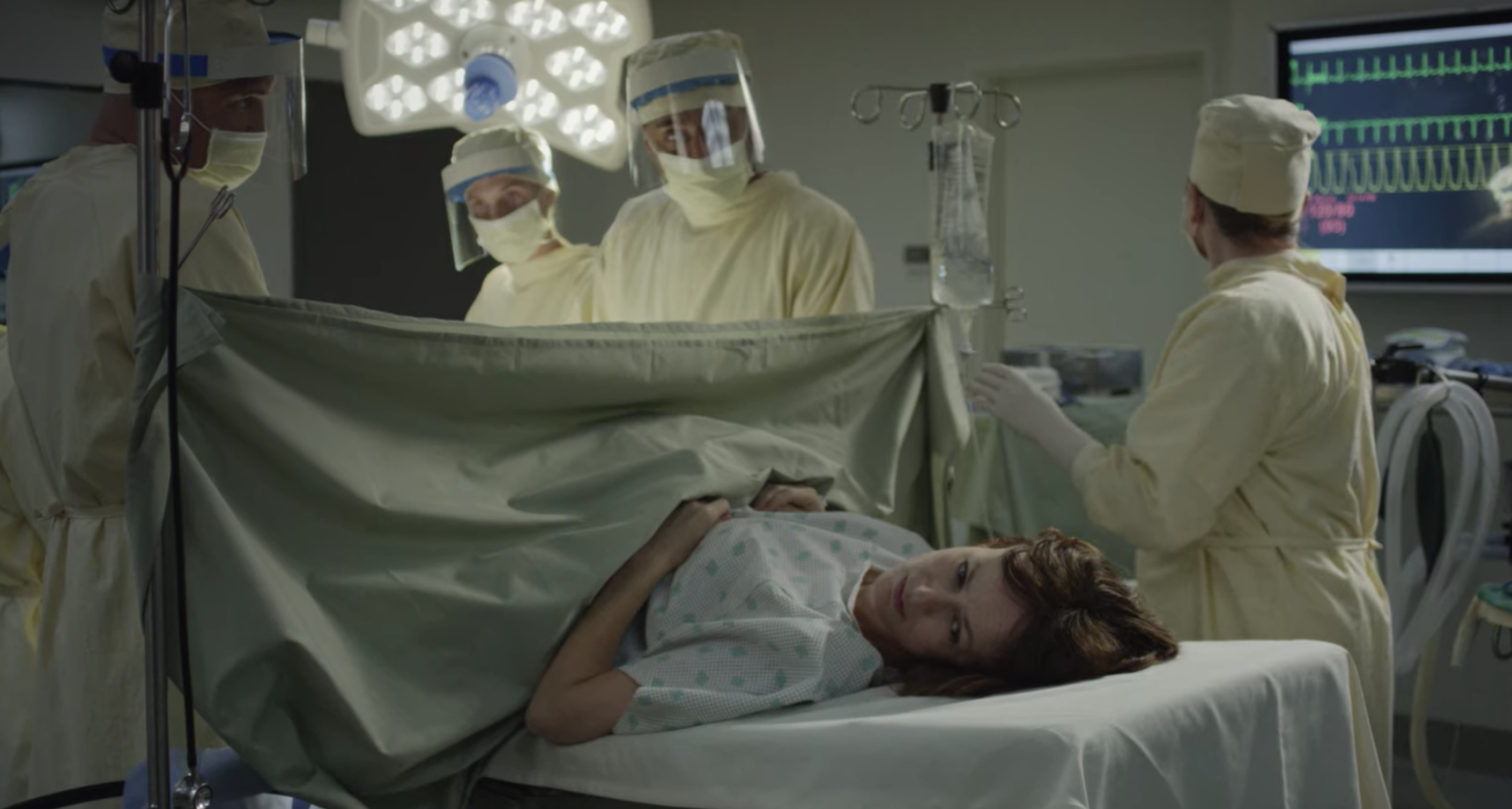 Woman lying on a hospital bed with a sheet held up below her chest as doctors give her a C-section