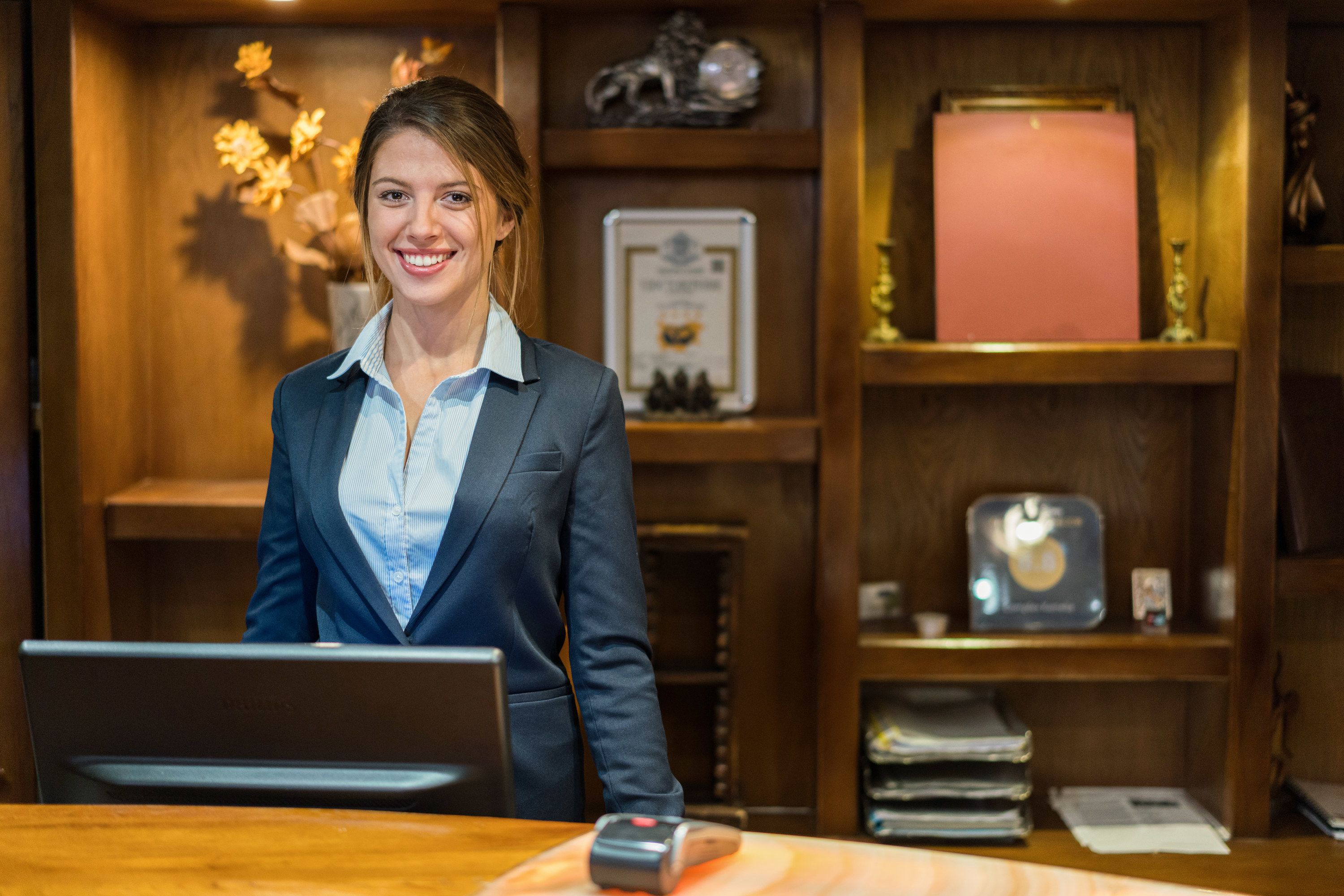 A woman standing at a hotel&#x27;s check-in desk smiling in a dark suit