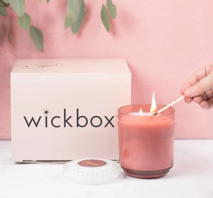A hand lighting a pink candle next to Wickbox&#x27;s packaging
