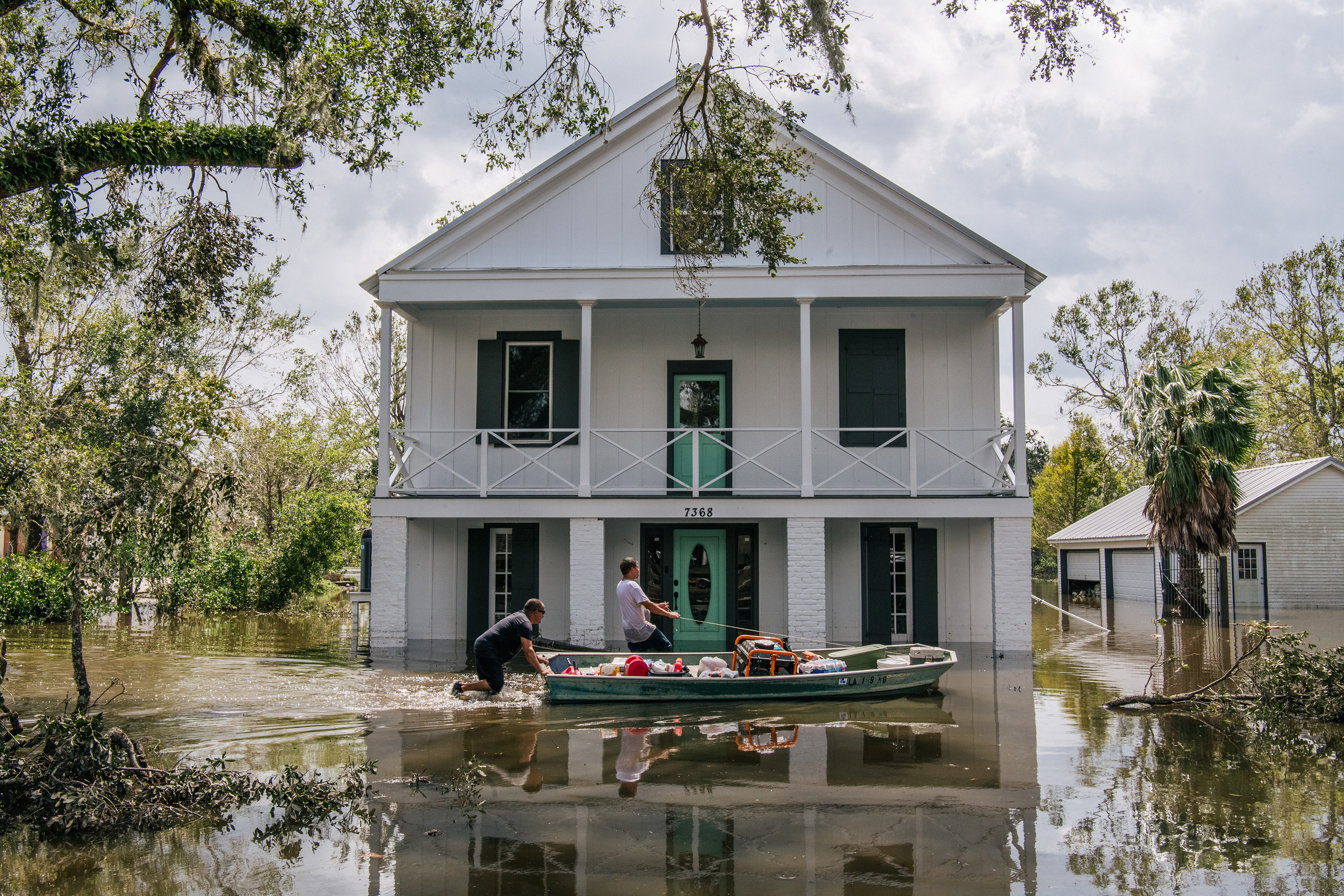 Two men maneuver a boat through water in front of a three-story house during flooding 