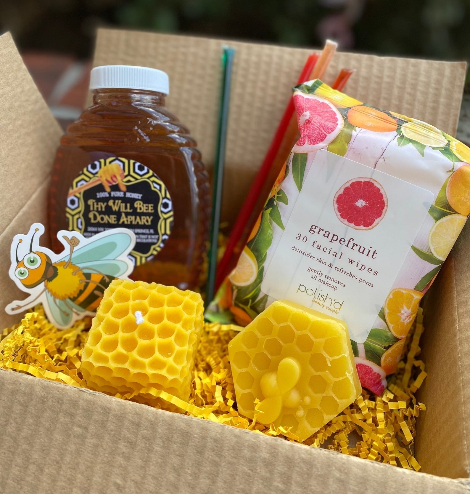 The Hive Life Bee Box containing a jar of honey, beeswax candles, facial wipes, and a bee sticker