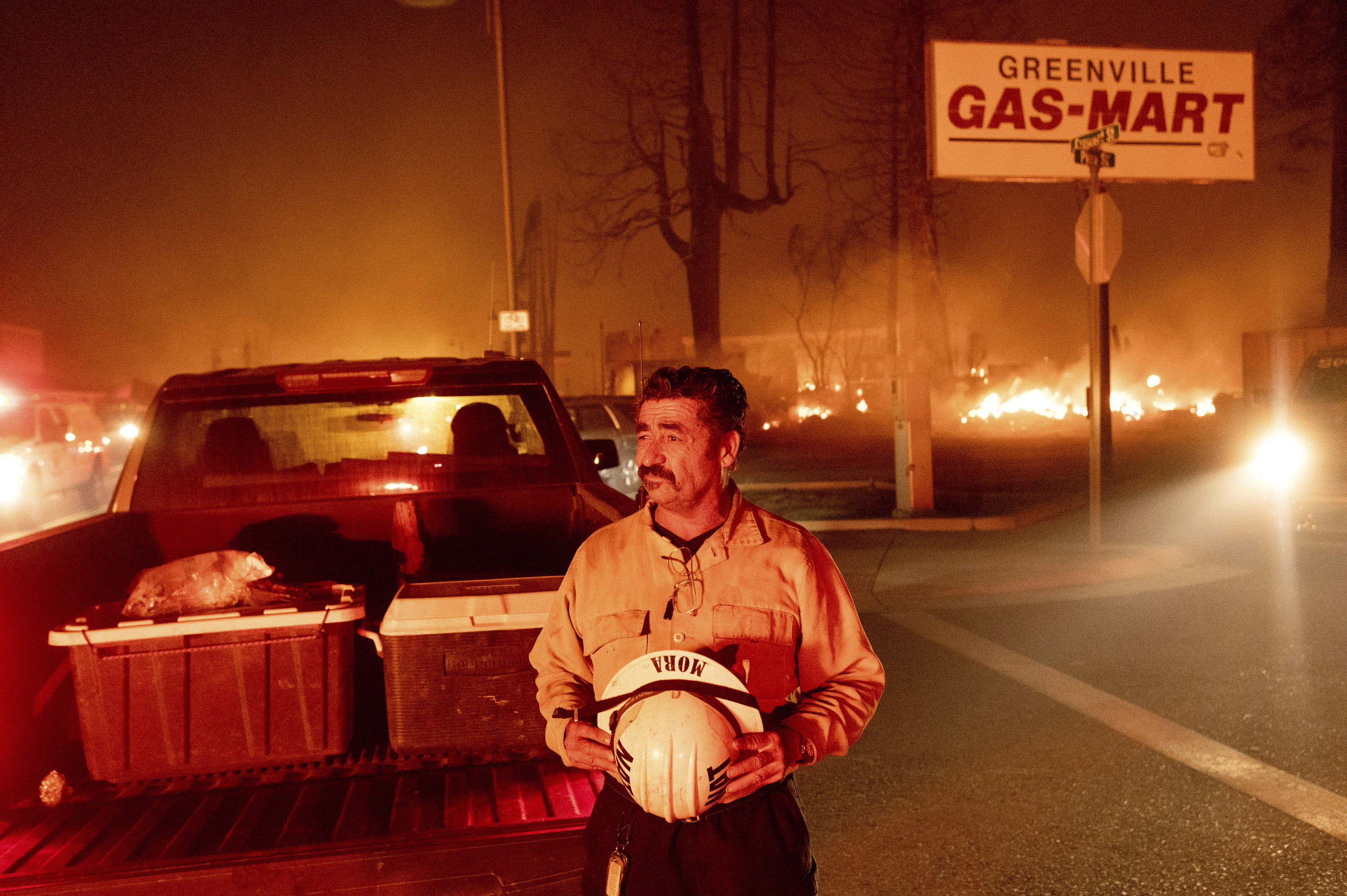 A man holds a helmet that says mora next to a truck with coolers and a fire raging behind him 