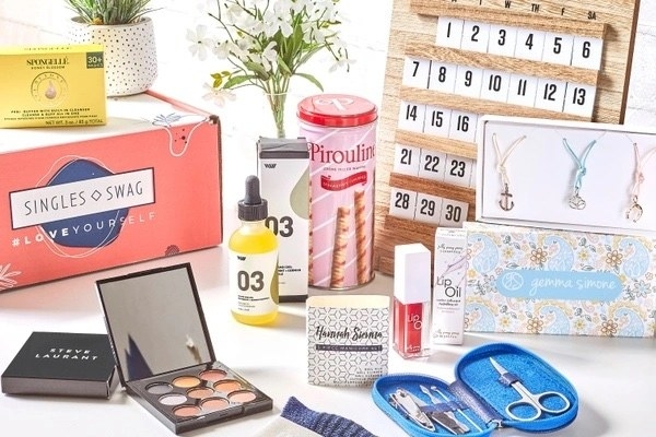 The SinglesSwag subscription box surrounded by its contents, including a makeup palette, cookie tin, skincare products, jewelry, and nail accessories