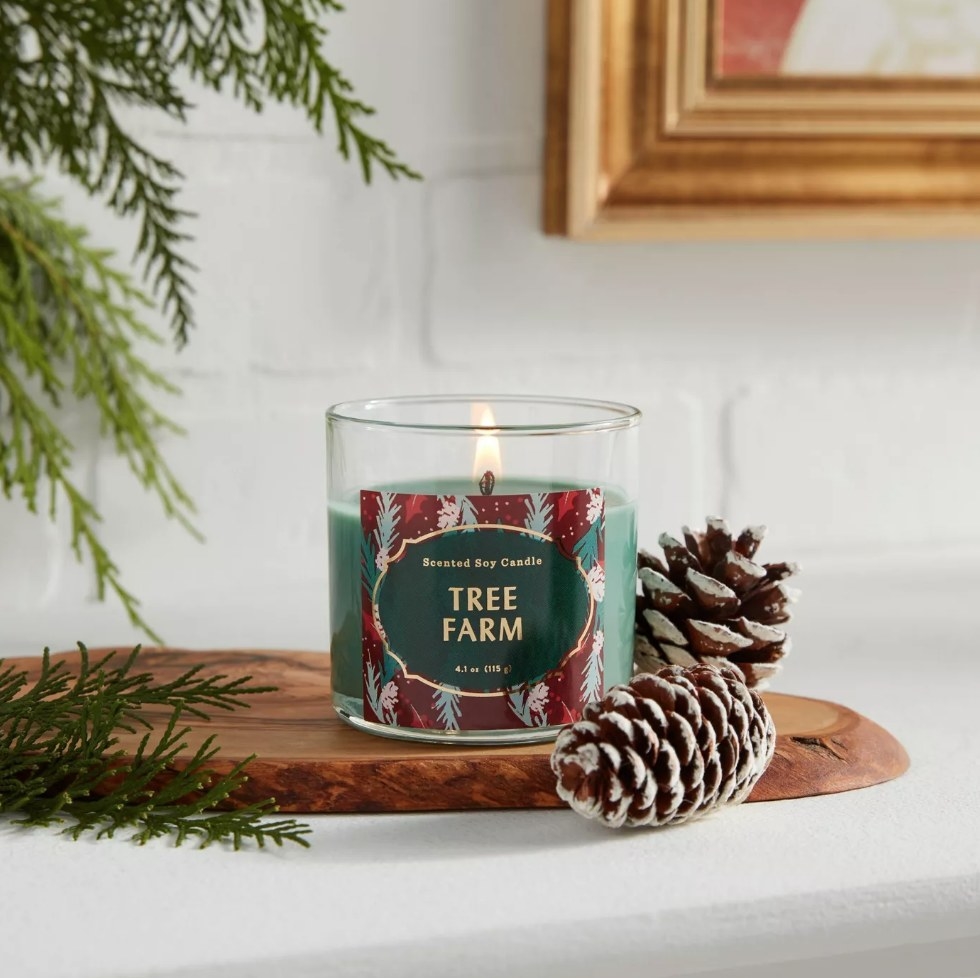 Green tree farm candle on wooden slab next to frosted pine cones