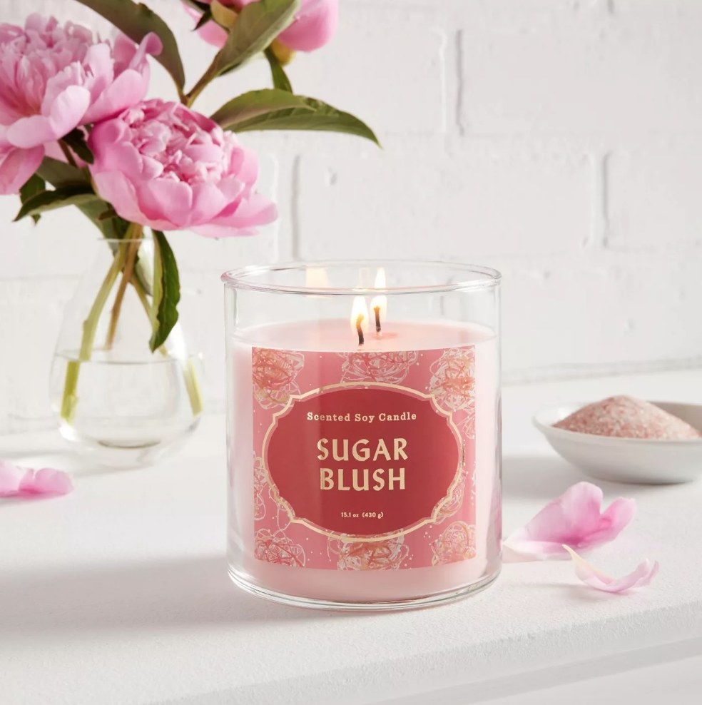 Pink candle with red sugar blush label sitting on white table next to pink flower petals, and pink flowers behind it