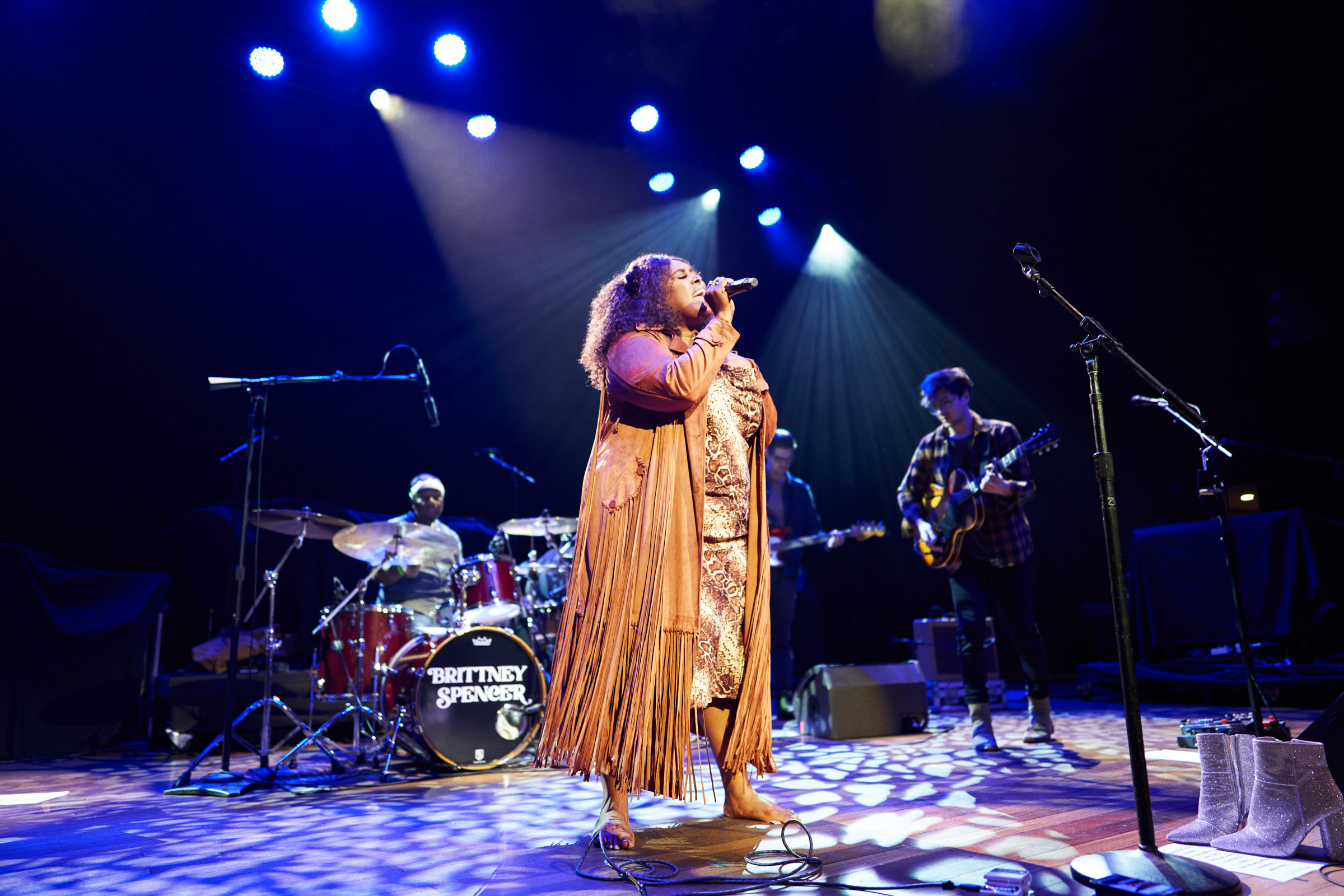 Four bandmates perform onstage with the singer, a Black woman wearing a full-length coat, is centered