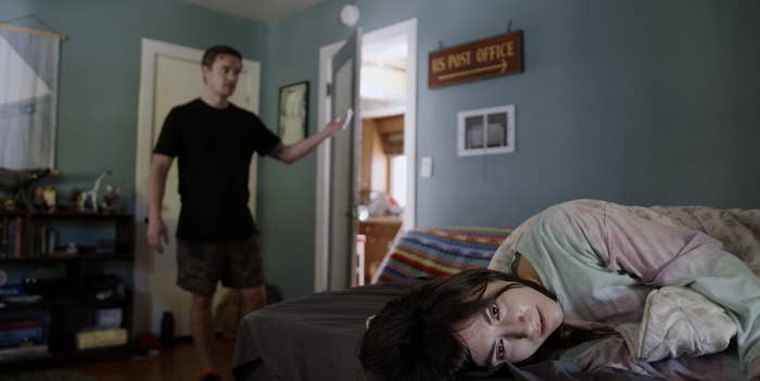 A woman lying on a bed staring blankly as a man stands near the door
