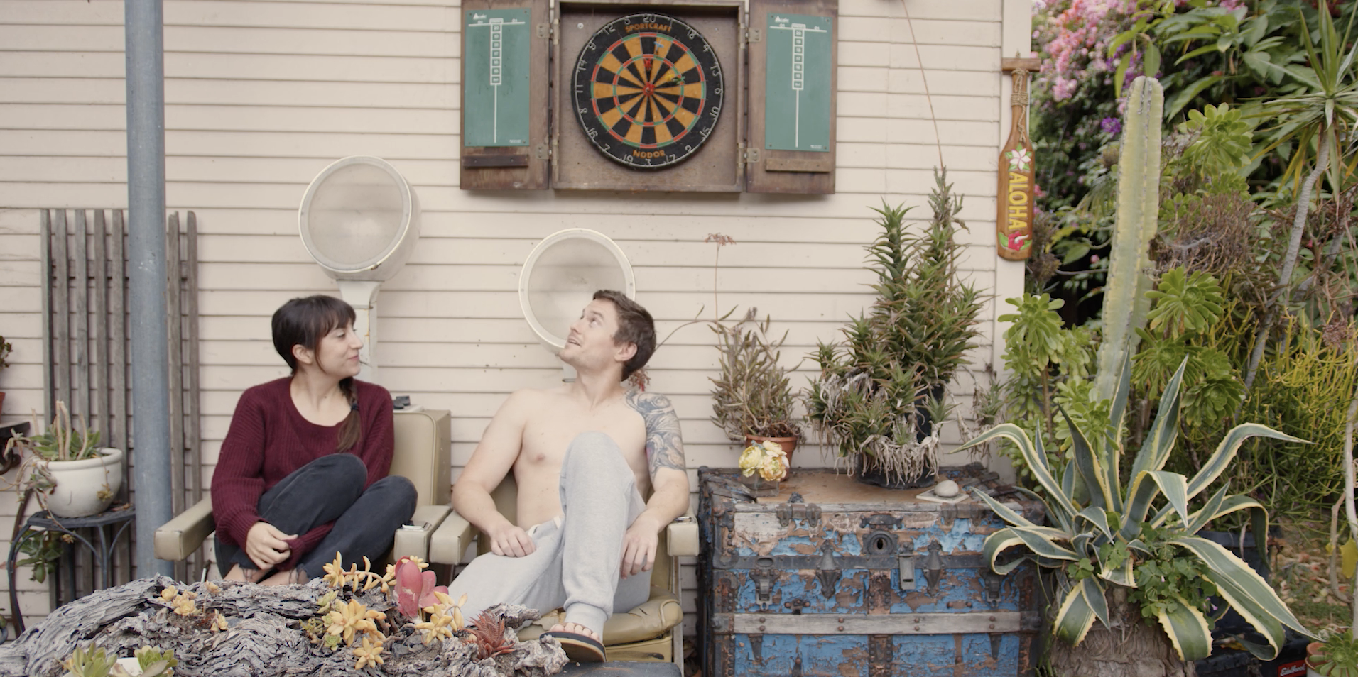 leah and nick sit in garden chairs in front of a small house with cactus next to them and a darts board above them