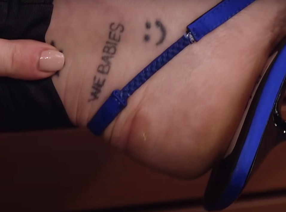 the words &quot;we babies&quot; on Miley&#x27;s ankle in all caps above a smiley face tattoo