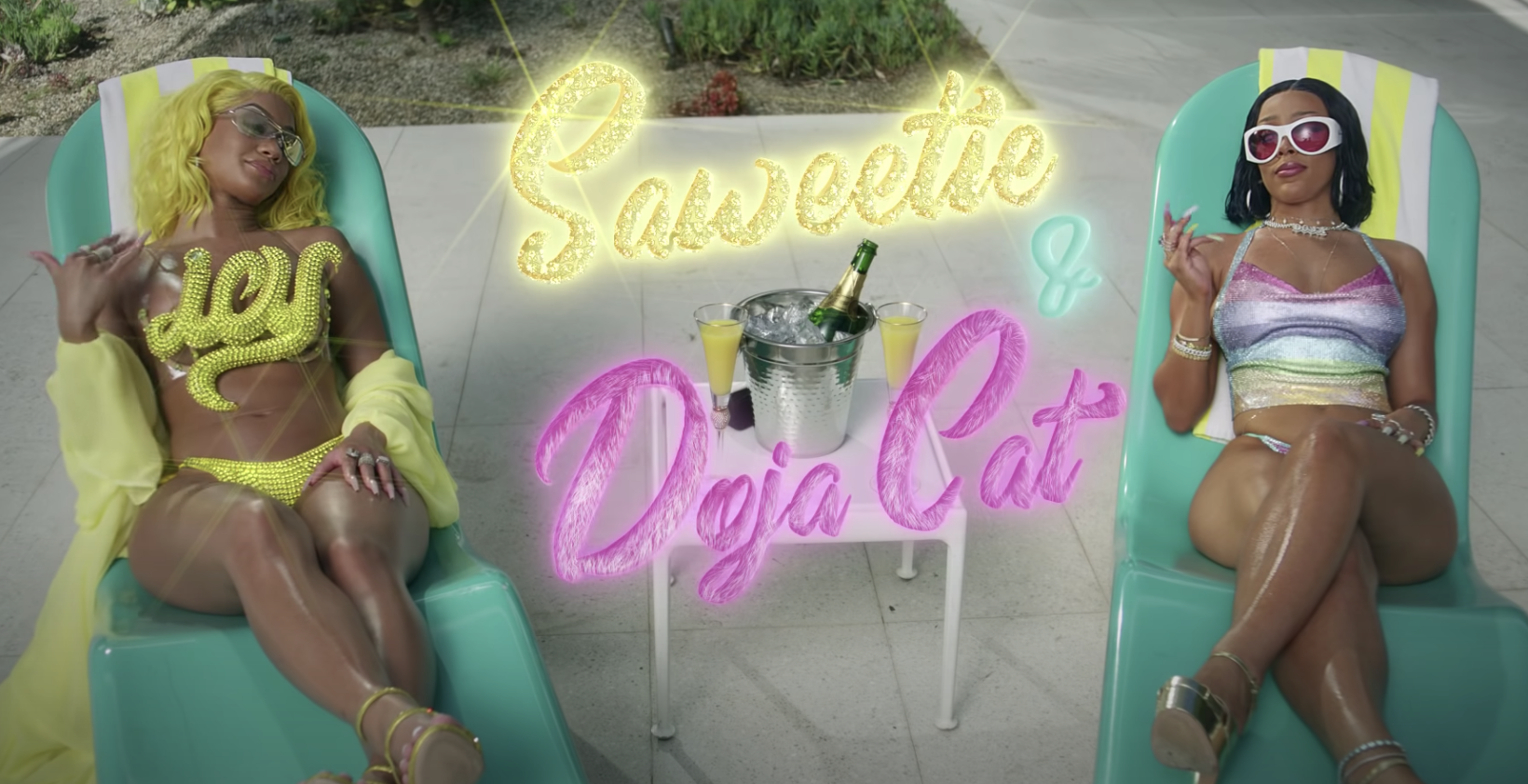 Saweetie and Doja Cat lounging on seats by the pool with Saweetie rocking a top that says ICY