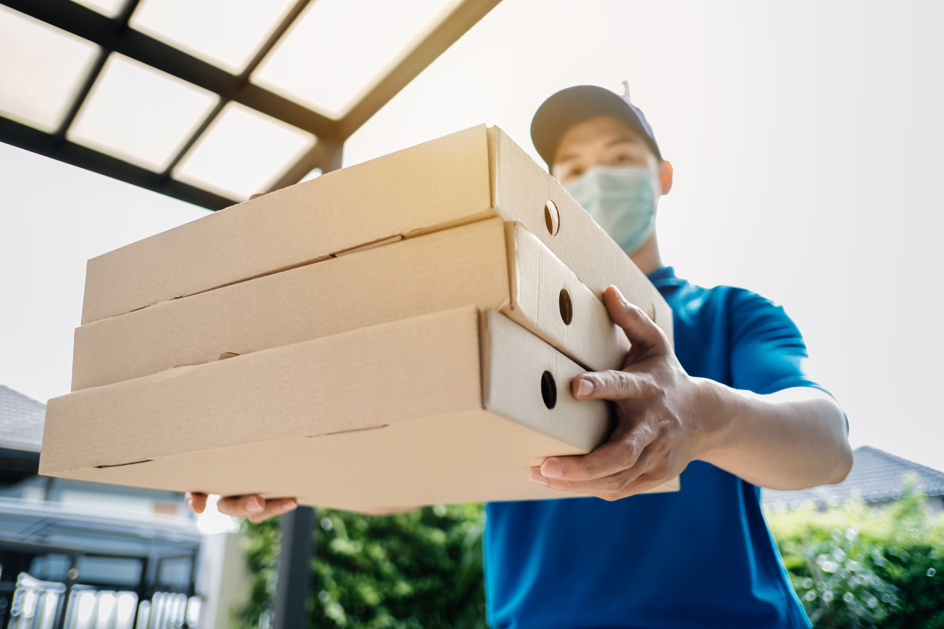 A pizza delivery man wearing a blue shirt and mask handing over three boxes of pizza