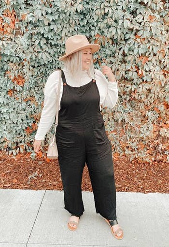 a reviewer wears the black jumpsuit with a shirt underneath and a beige hat