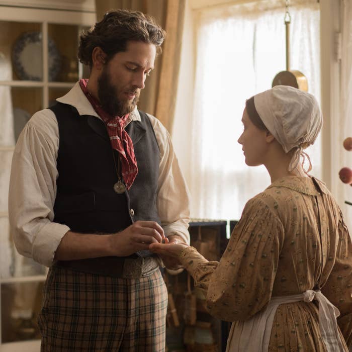 A bearded man wearing a black vest and red scarf looks down at a woman's hand he's holding in his hand. The woman looks at him.