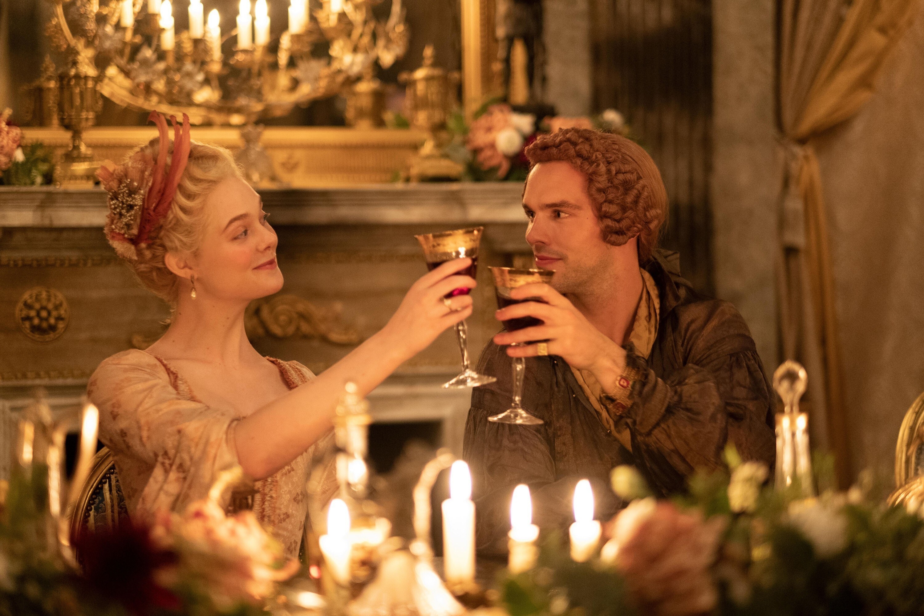 A blonde woman and a man with a curly wig cheers with glasses of red wine. In front of them are candles.
