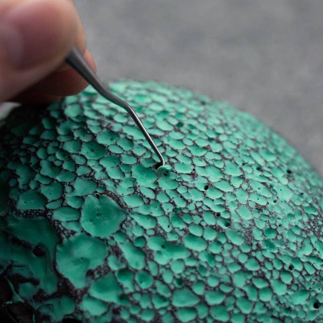 a hand using a scraping tool to remove blue latex from the holes in a lava stone