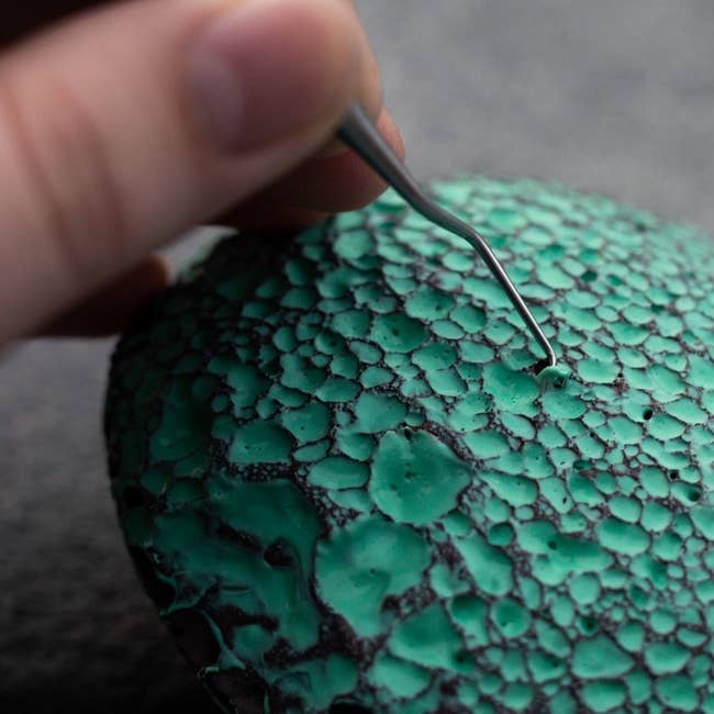a hand using a scraping tool to remove blue latex from the holes in a lava stone