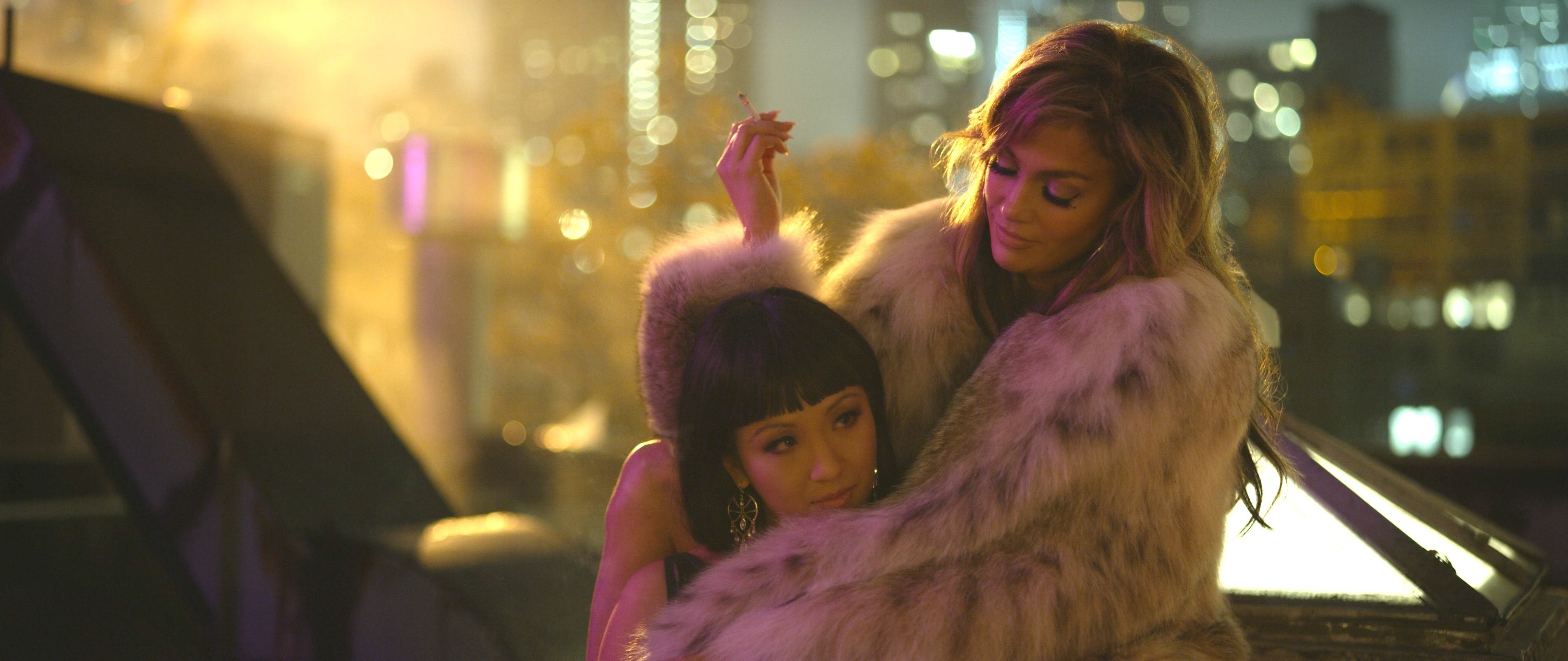 Constance Wu and Jennifer Lopez wrapped in a fur coat on a roof top