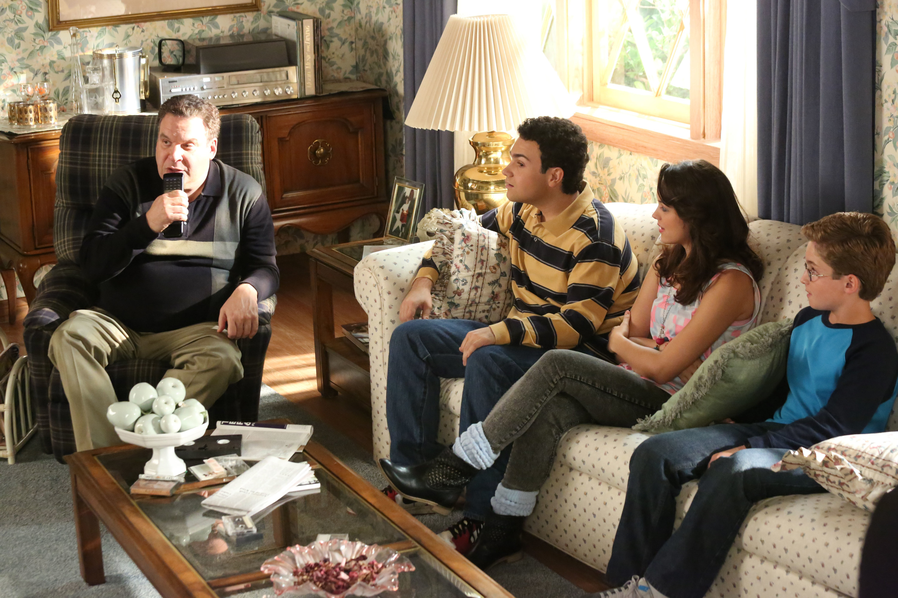 Garlin puts a remote control to his chin as he sits in a living room with his kids in a scene from the show