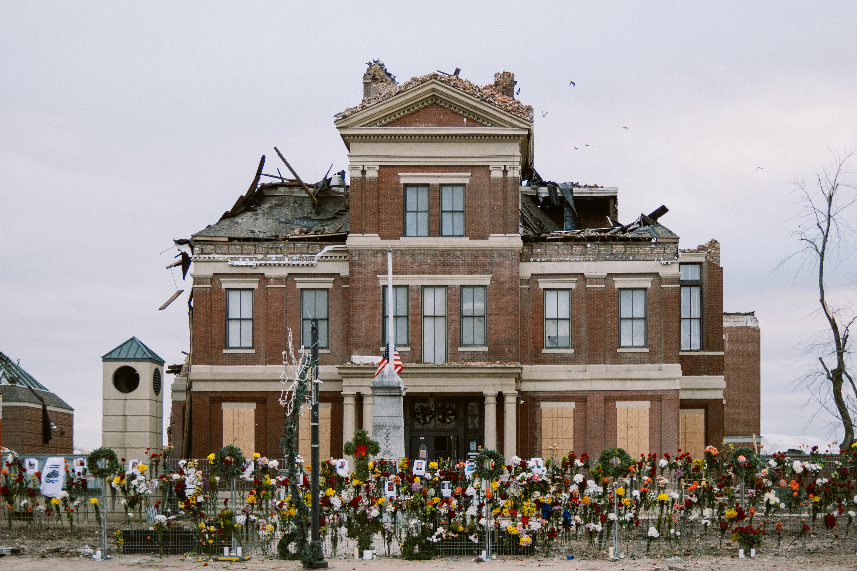 A damaged courthouse with the roof missing in parts, the flag down, and flowers and wreaths along the fence 