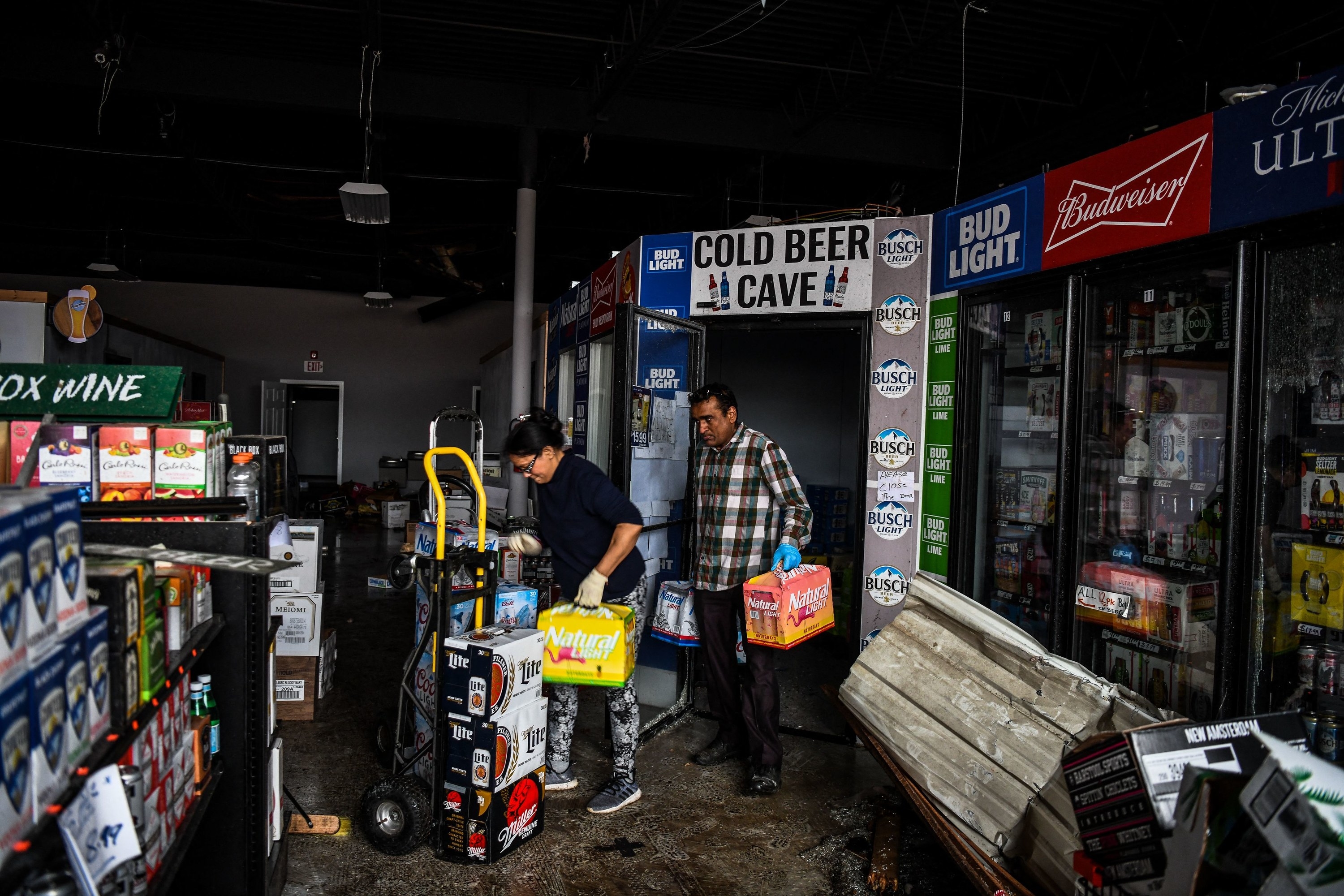 Two people load beer out from a cooler in a destroyed liquor store