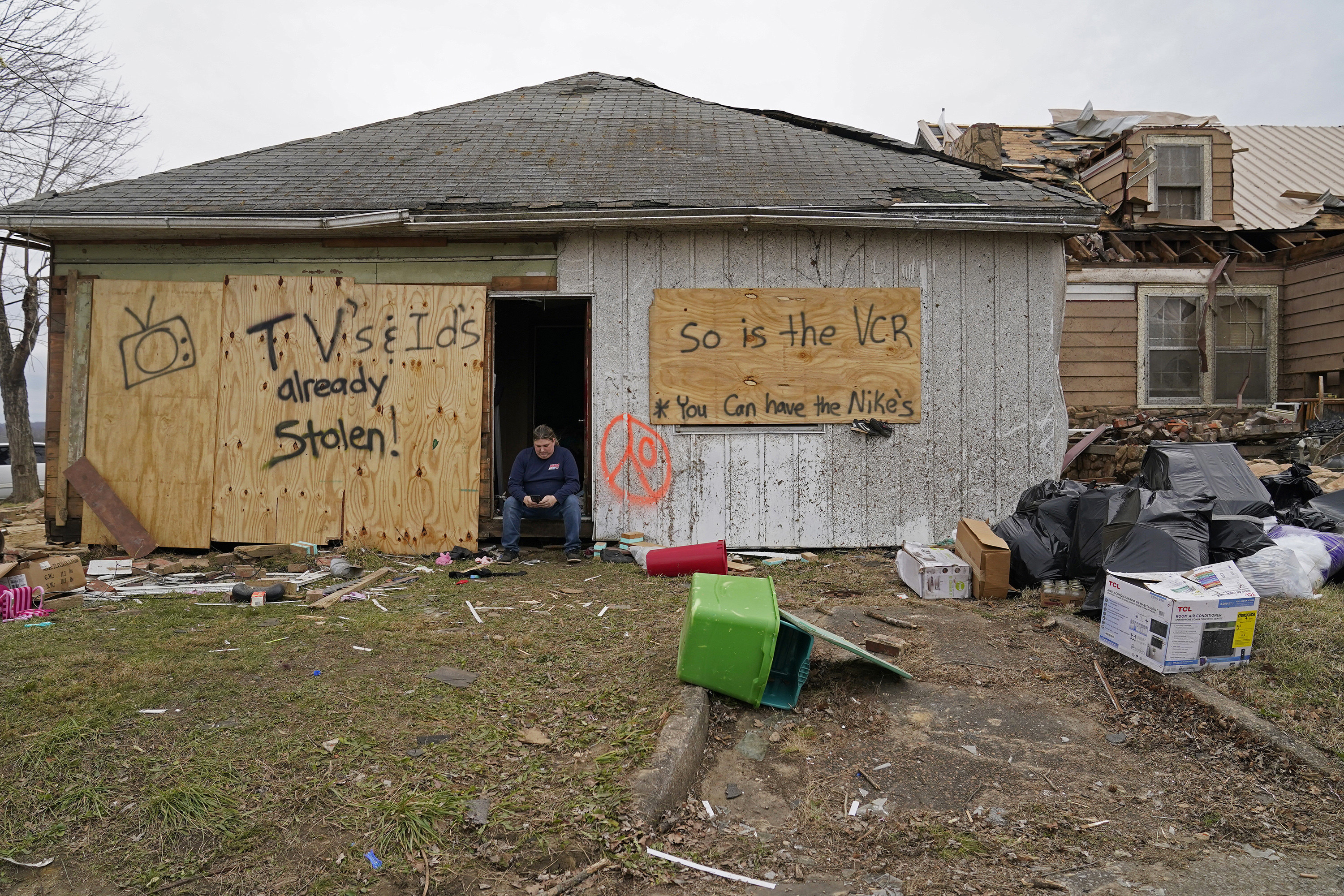 A man sits in the doorway of a destroyed home with signs that read tvs and ids already stolen so is the vcr you can have the nikes