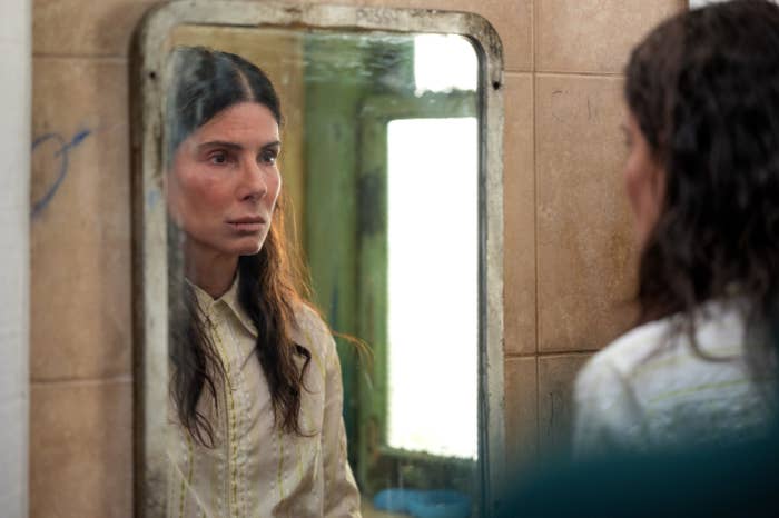 Woman looks blankly into a mirror shrouded by beige tile