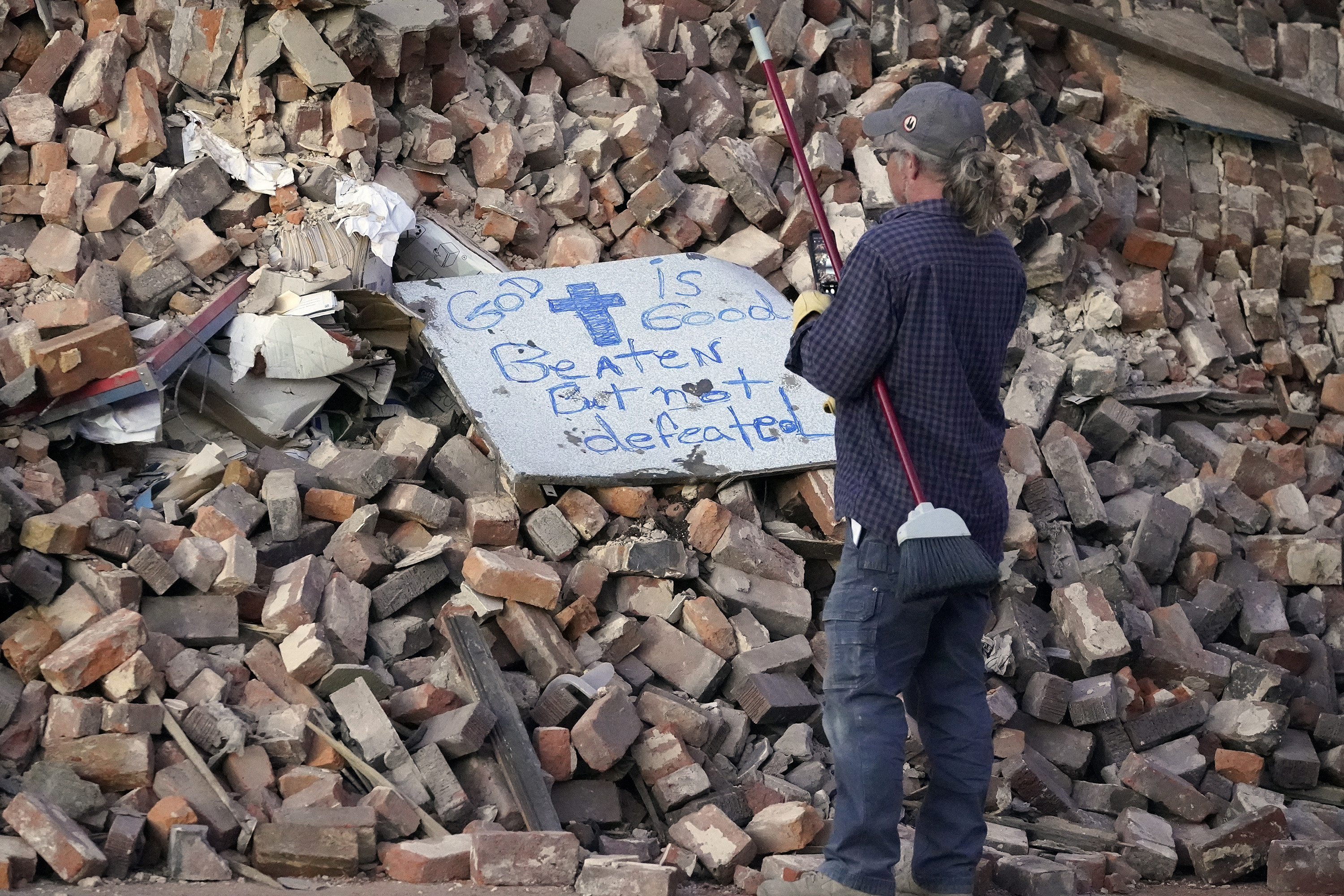 A man with a broom takes a photo on a pile of bricks that says that god is good beaten but not defeated