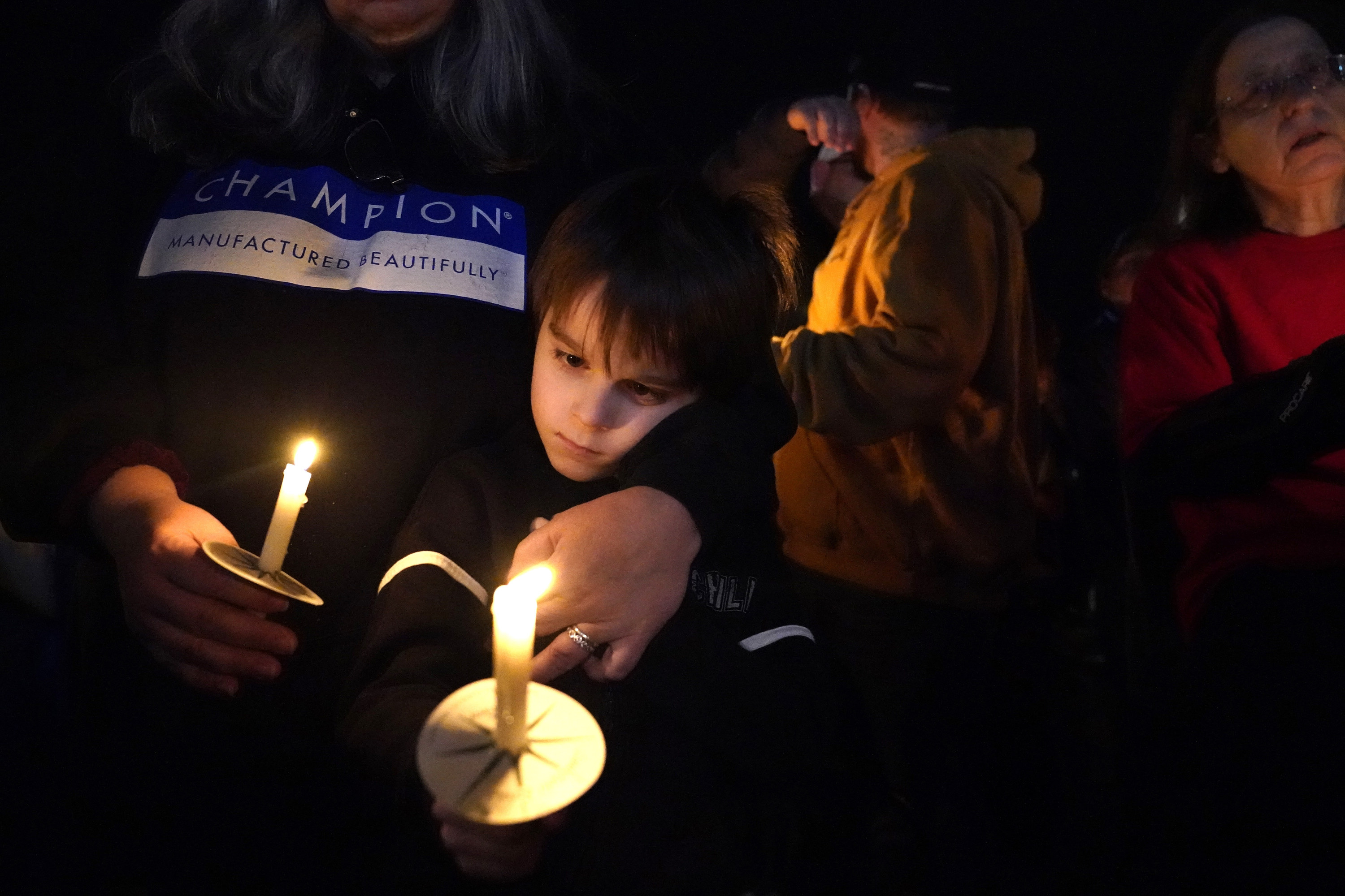 A young boy and his mother with two candles during a vigil, people behind them 