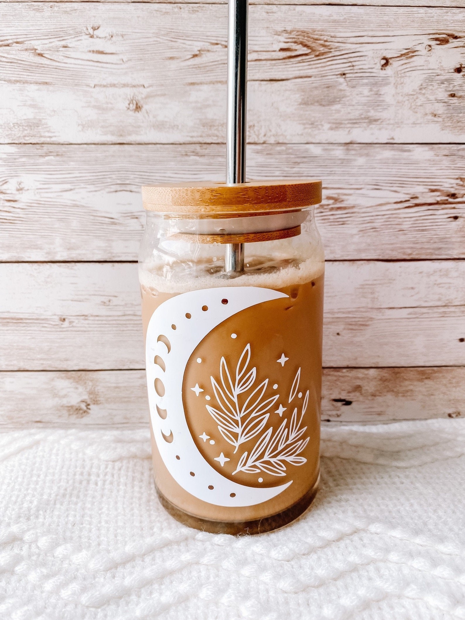 A glass cup with iced coffee inside and a decal of a moon and leaves on the front