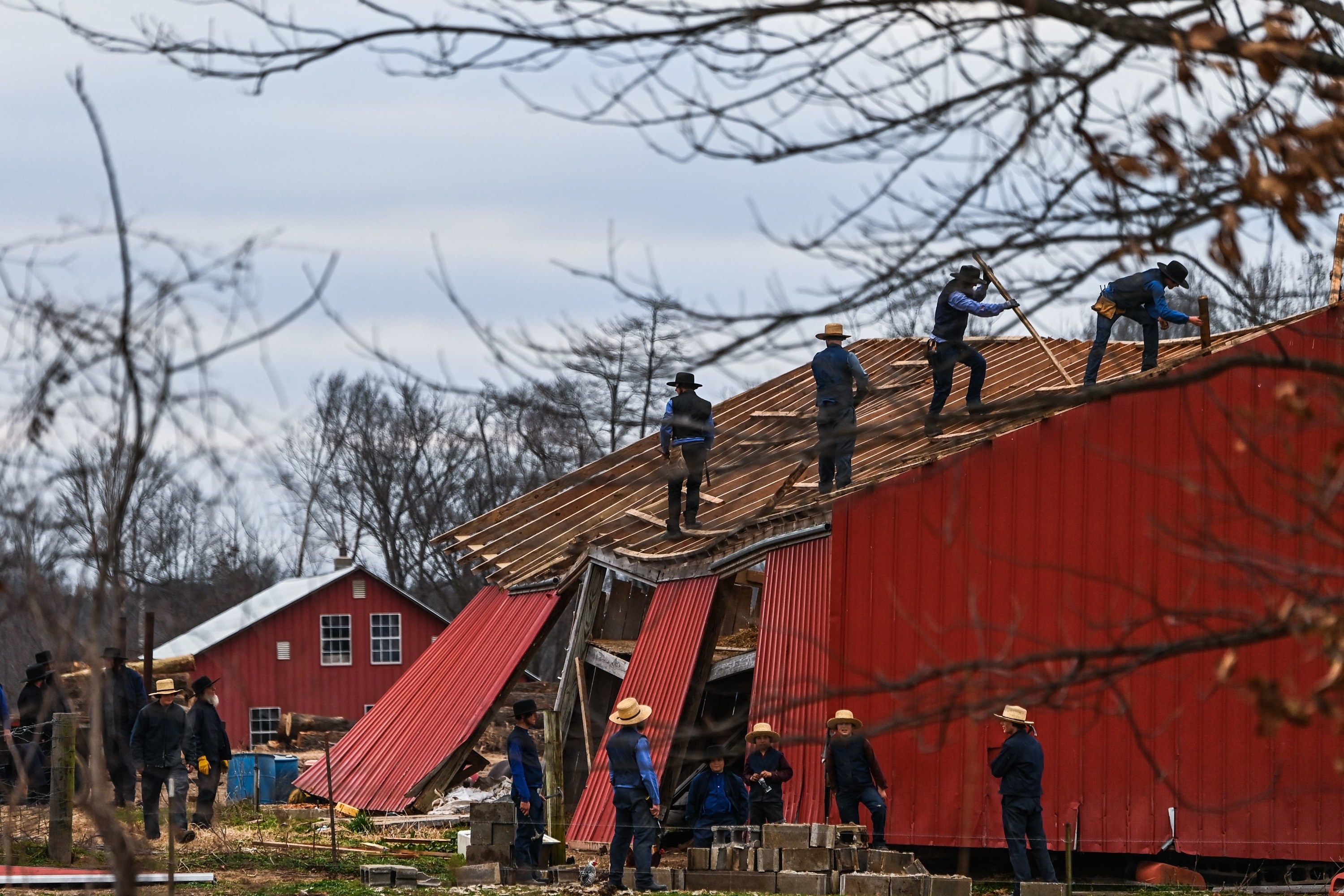 Workers on top and below a barn try to repair it, all wearing denim and Amish hats