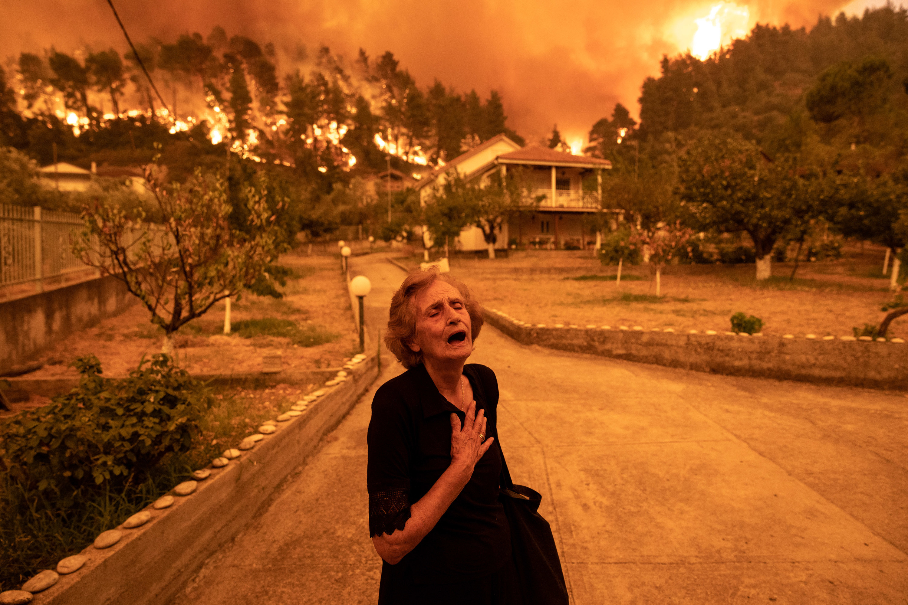 An older woman dressed in black clutches her chest while the landscape behind her burns orange