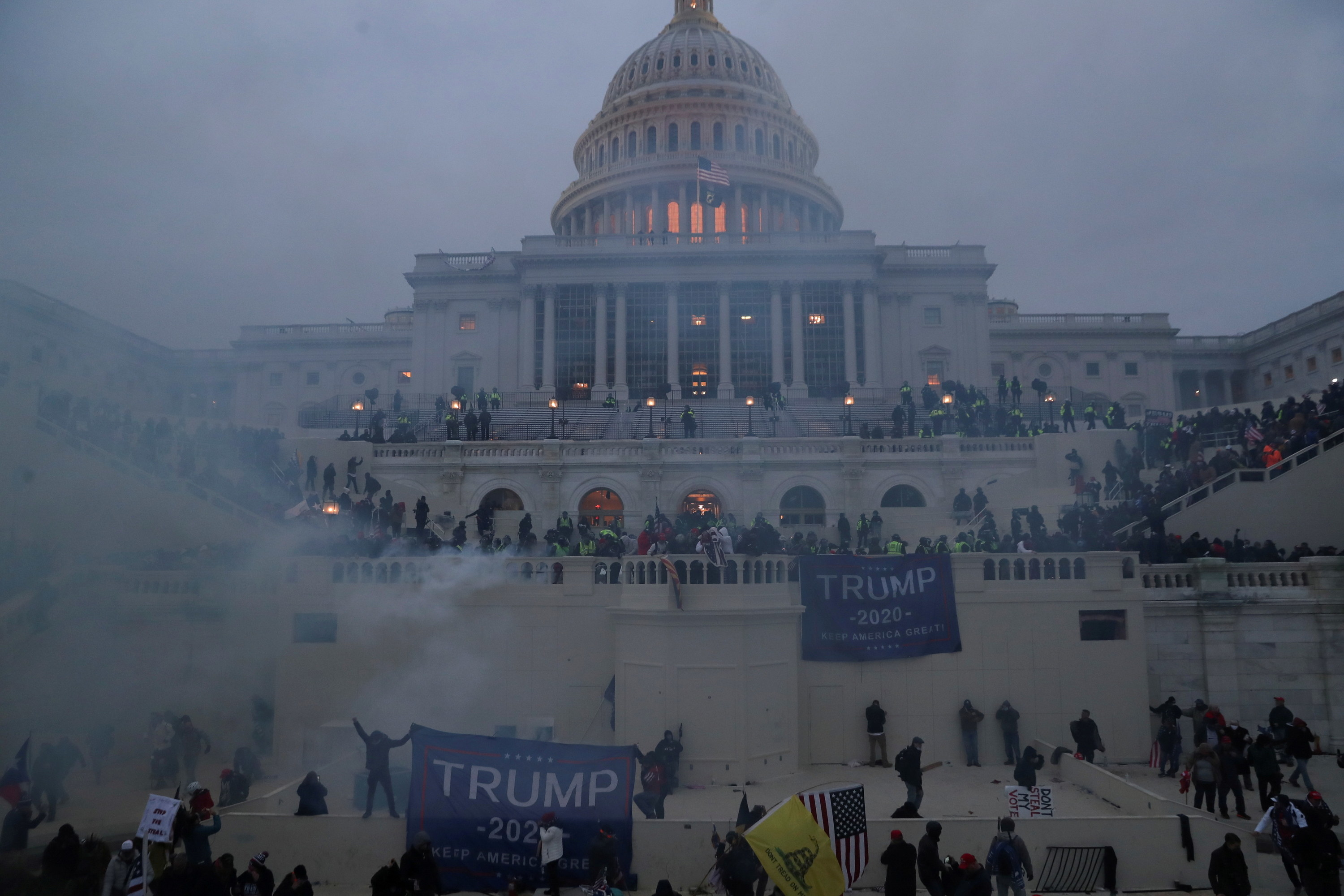 The capitol building, with lights on inside, is seen through smoke as hundreds of police and protesters carrying trump signs are on the steps in a chaotic scene 