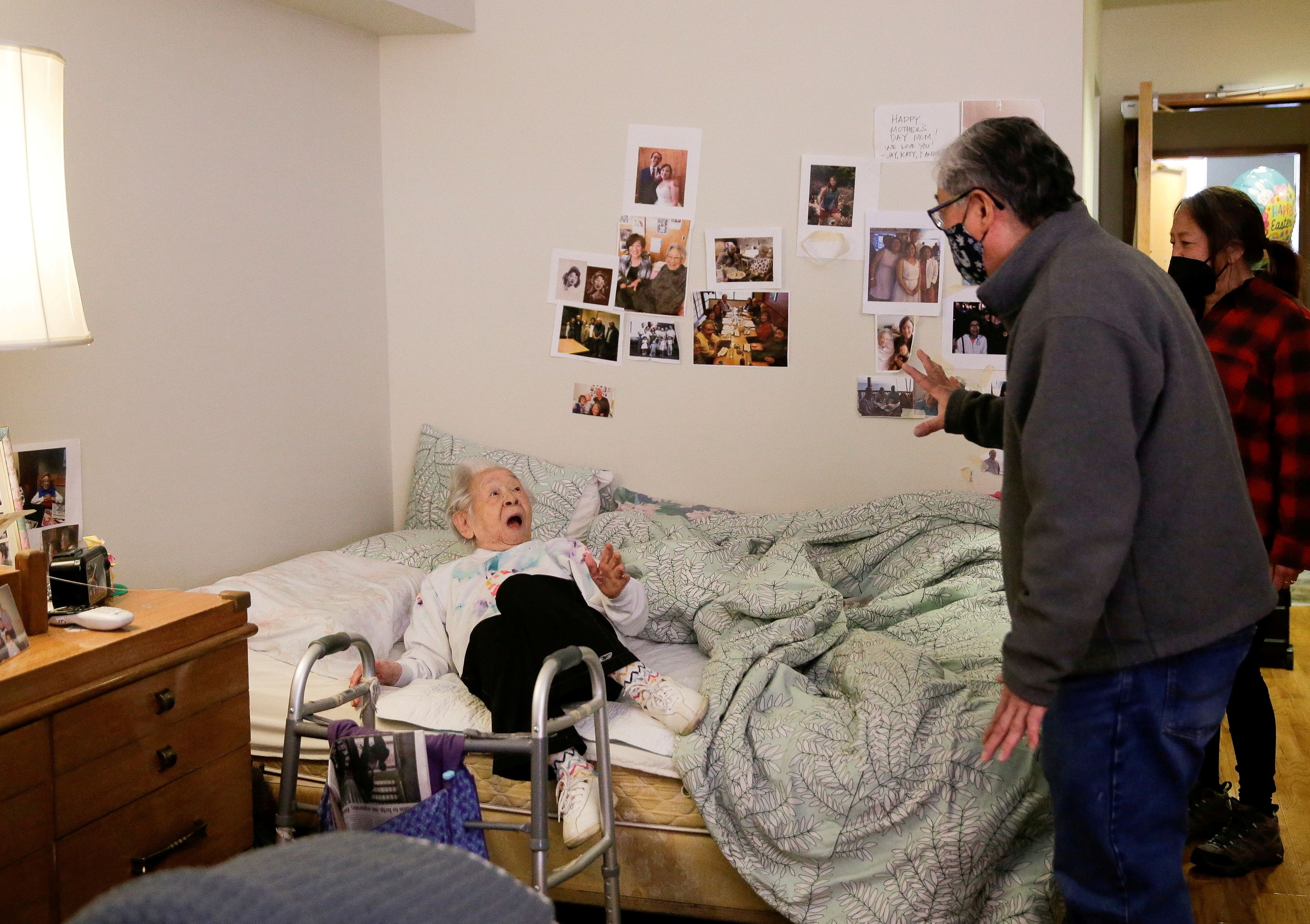 An older woman on a bed looks shocked as a man enters her room with a woman