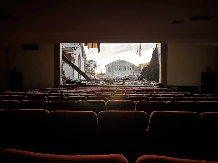 Several rows of seats in a movie theater sit empty; the wall where the screen would be is missing, revealing fallen debris from a tornado