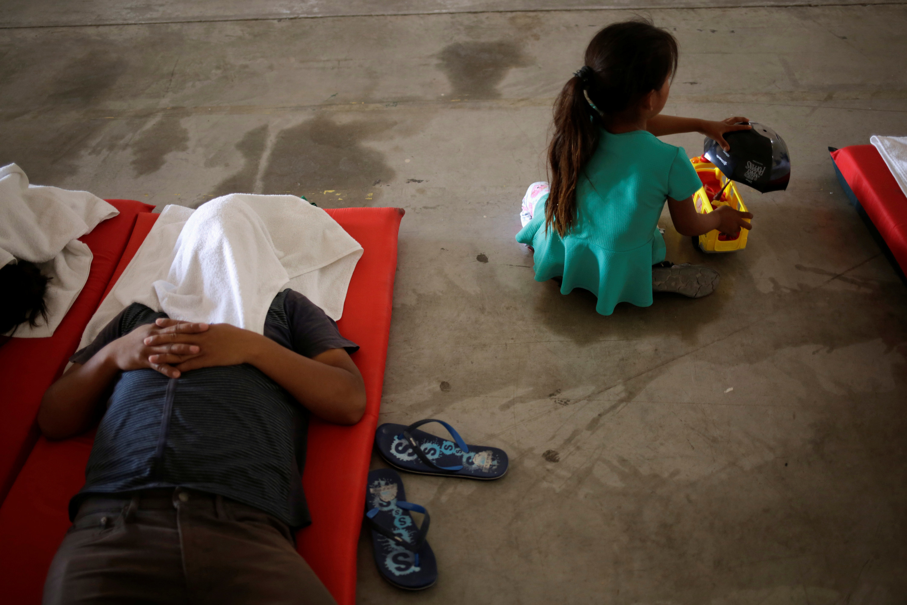 A little girl sits on the floor, and a person lies on a cot with a cloth over their face