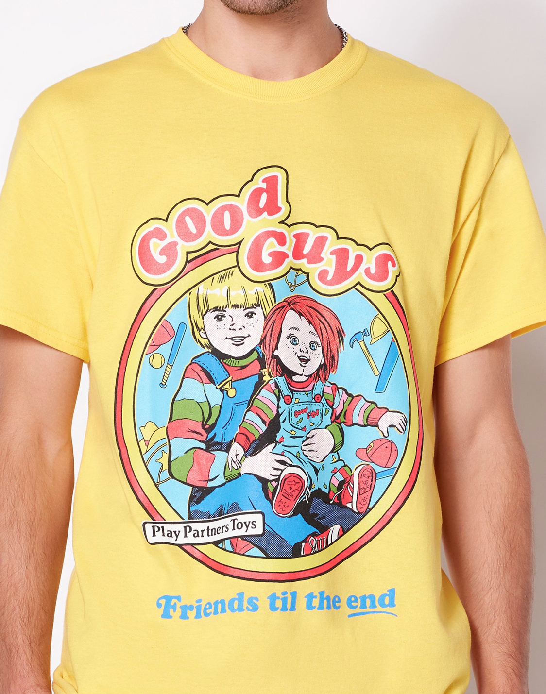 A yellow t shirt with a vintage logo that says good guys and shows a kid in overalls holding a chucky doll and says Friends til the end at the bottom